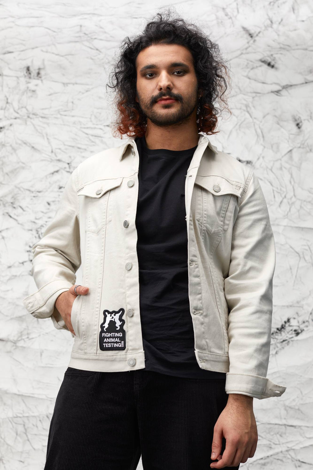 Model is stood in front of a marbled background wearing all black with a white denim jacket with the patch on the bottom.
