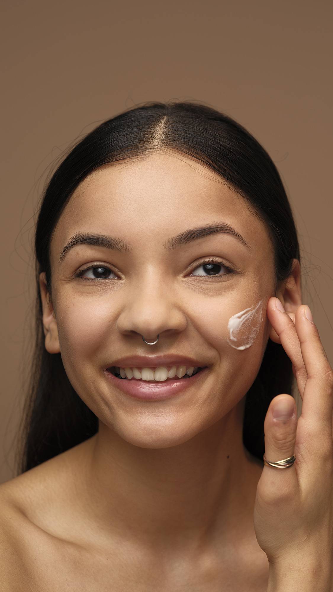 A model with long dark hair and a septum piercing smiles broadly while dabbing a patch of white Enzymion face cream onto their cheek. They are shown from the shoulders up against a solid brown background.