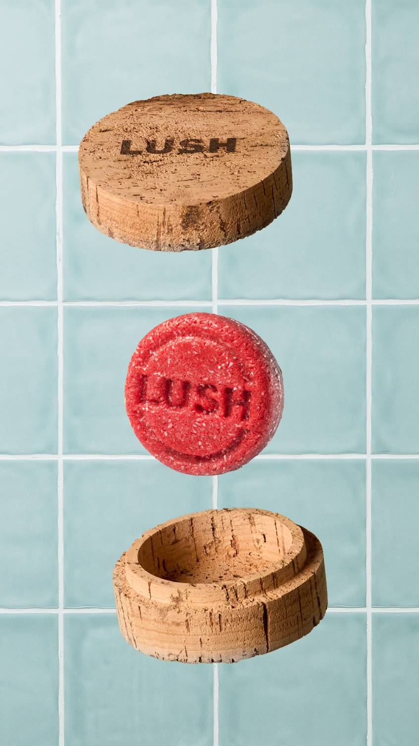 A red circular Lush "NEW" shampoo bar floats in the centre, positioned between the lid and base of an open, round cork pot. The lid is branded with the 'LUSH' logo. Set against a background of pale blue-green bathroom tiles.