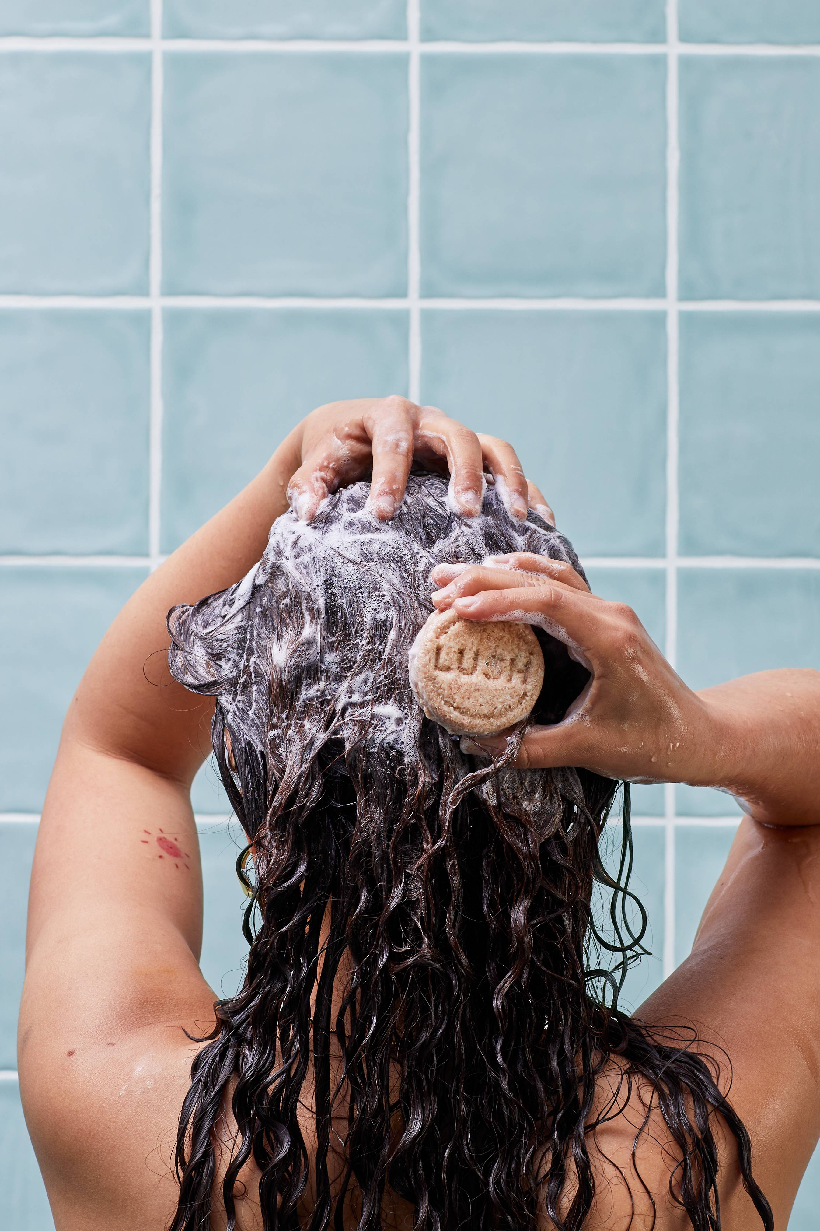 A smiling person in a shower washes their long, wet dark hair. They hold a round, beige Lush solid shampoo bar, lathering it into their wavy locks. The background features light teal-blue square wall tiles with white grout.