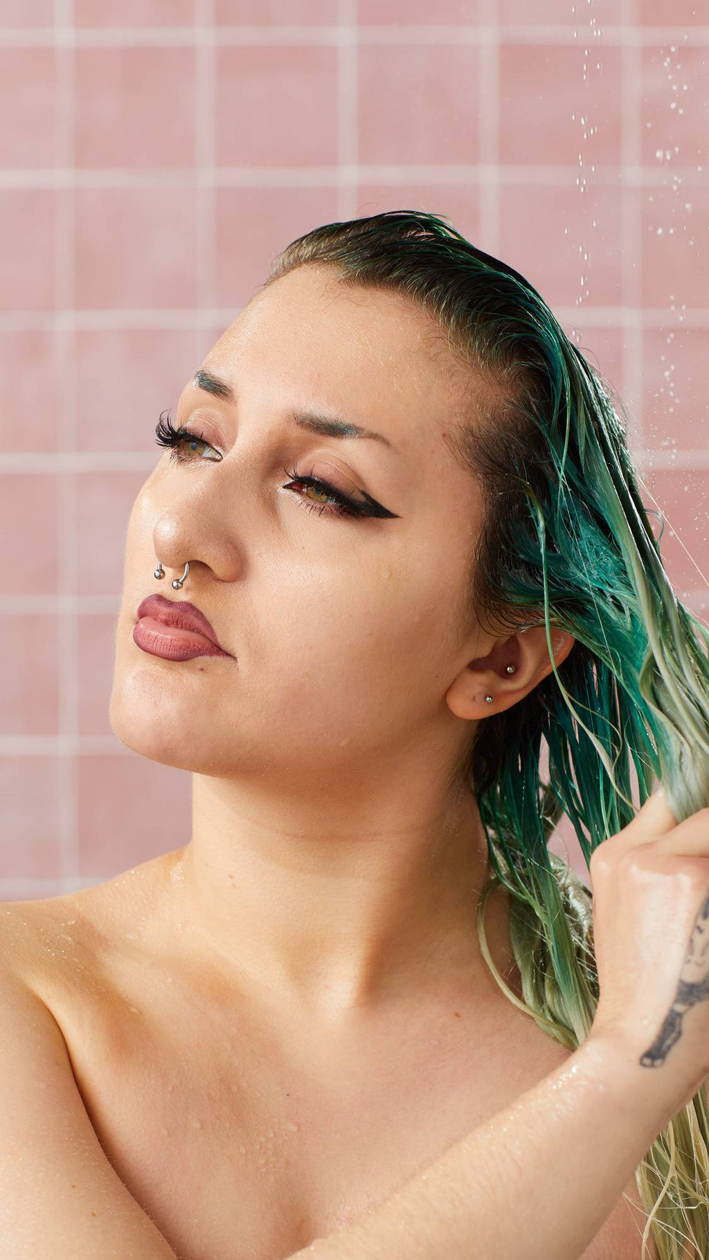 A model with winged eyeliner and facial piercings washes their wet, green-tinted hair in the shower with the Lush "Retread" conditioner. Water droplets fall around them against a background of pink square tiles.
