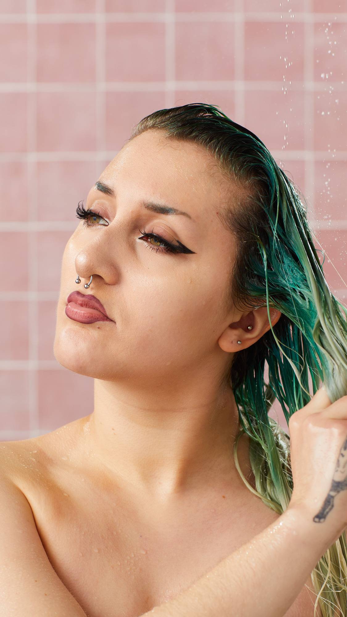 A model with winged eyeliner and facial piercings washes their wet, green-tinted hair in the shower with the Lush "Retread" conditioner. Water droplets fall around them against a background of pink square tiles.
