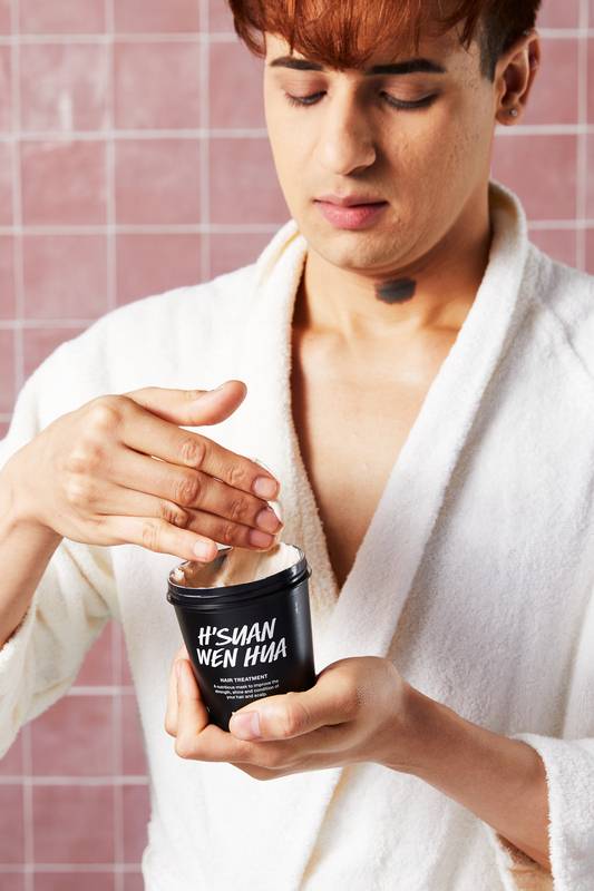 A model with copper-toned hair, wearing a white robe, holds a black tub of Lush 'H’suan Wen Hua' hair treatment. They are dipping their fingers into the creamy product against a background of pink square tiles.