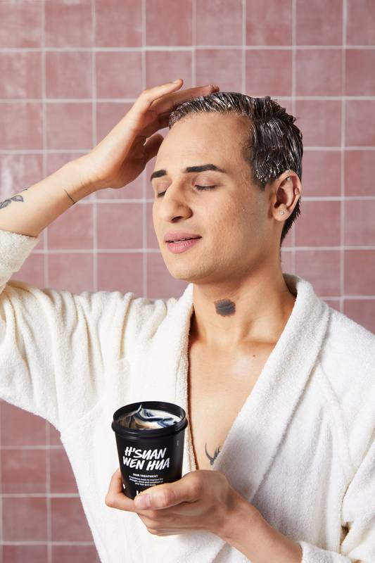 A model wearing a white robe massages a creamy hair treatment into their hair. They hold a black tub of Lush 'H’suan Wen Hua' in one hand, with their eyes closed in a relaxed expression against a pink-tiled wall.