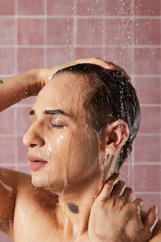 A model rinses the "H'suan Wen Hua" hair treatment out of their hair under a running shower. Their eyes are closed and they have their hands on their head and neck, appearing relaxed. The background consists of pink square wall tiles.