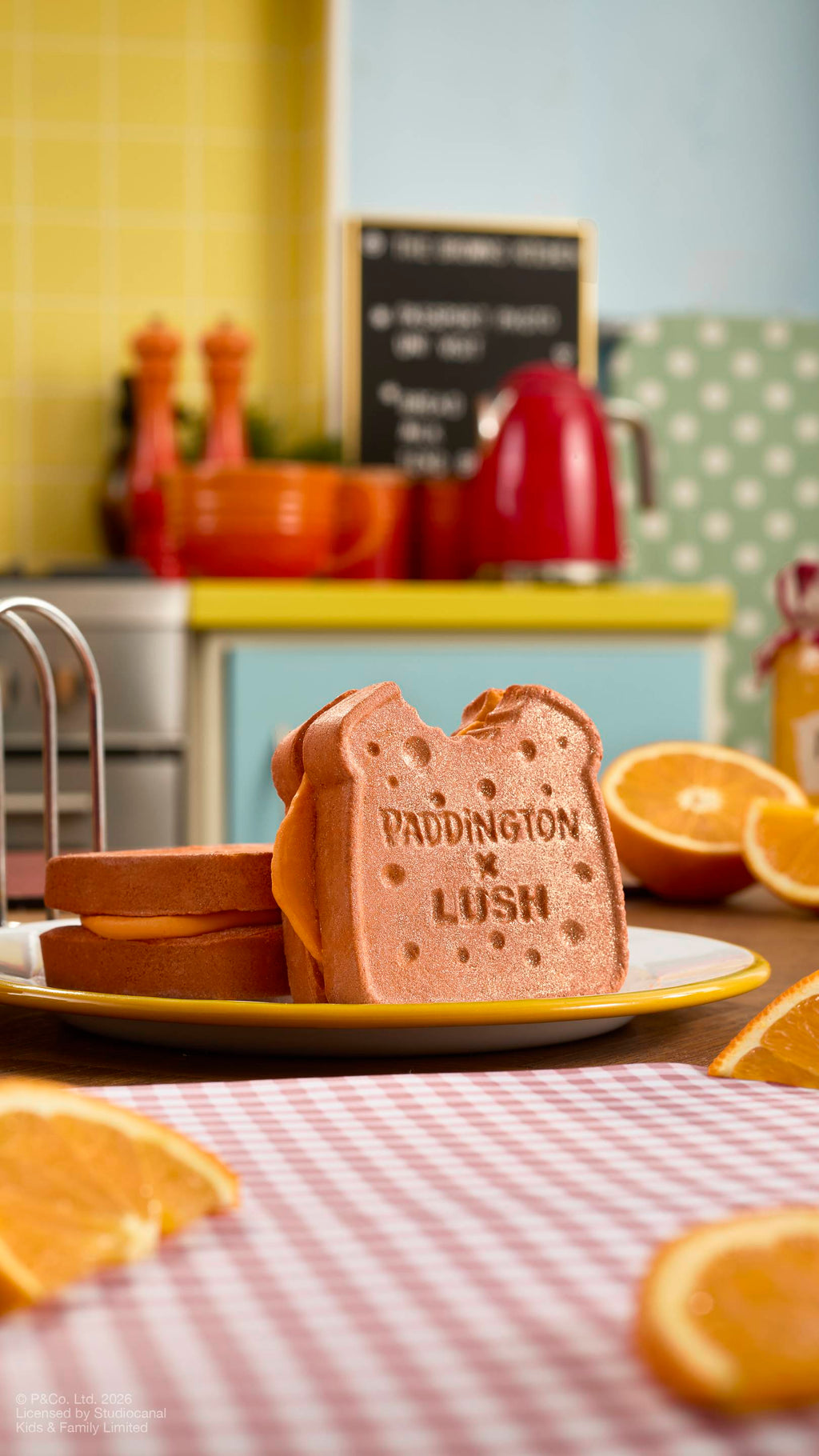 The image shows two brown, "Marmalade Sandwich" bubbleroon bubble bars sitting on a plate on a kitchen counter. One stands upright, showing a 'bite' taken out of the corner and 'PADDINGTON x LUSH' text. Surrounded by orange slices.