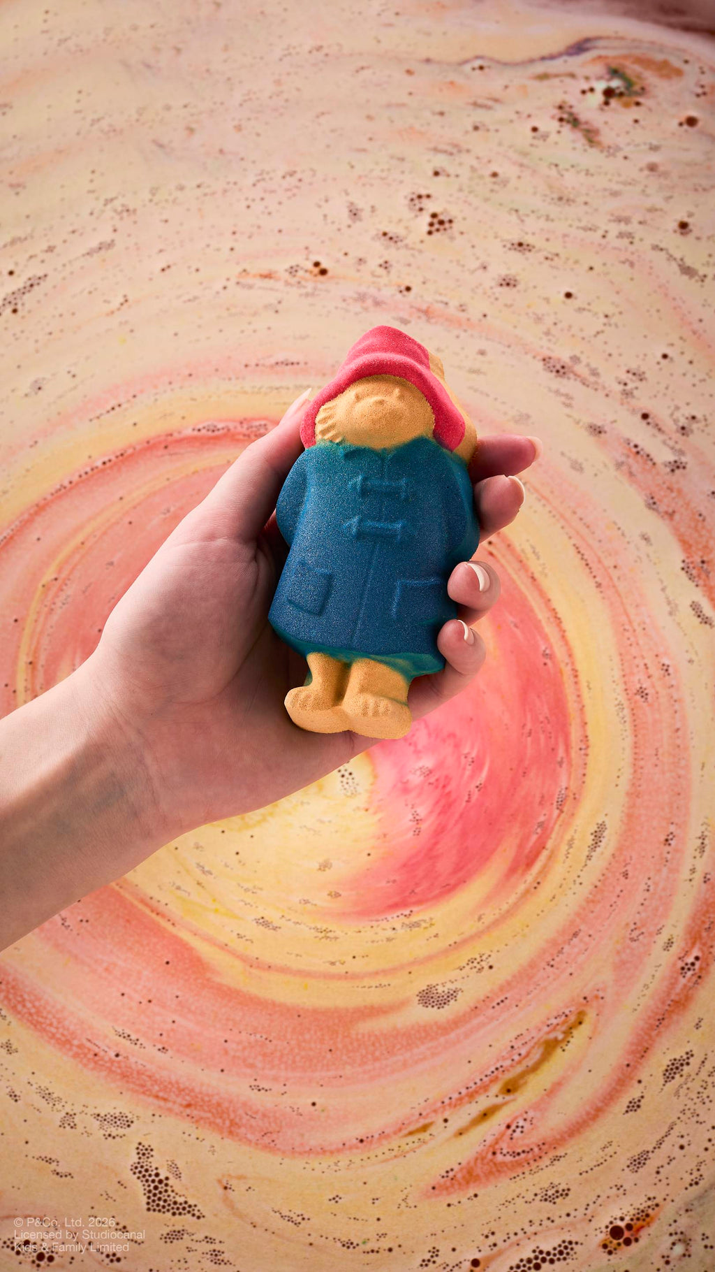 A model's hand holds a "Paddington Bear" bath bomb over bathwater. The bear is detailed with his signature blue coat and red hat. The background water shows a swirling whirlpool of peach, gold, and pink foam from a dissolving product.