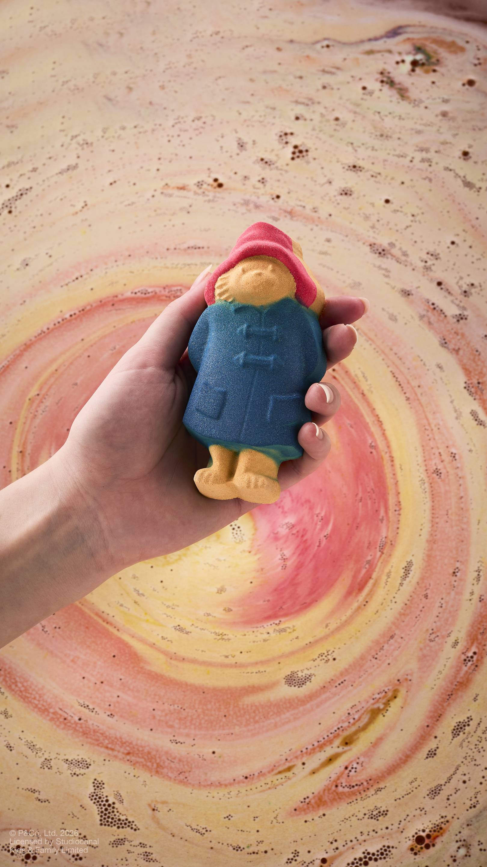 A model's hand holds a "Paddington Bear" bath bomb over bathwater. The bear is detailed with his signature blue coat and red hat. The background water shows a swirling whirlpool of peach, gold, and pink foam from a dissolving product.