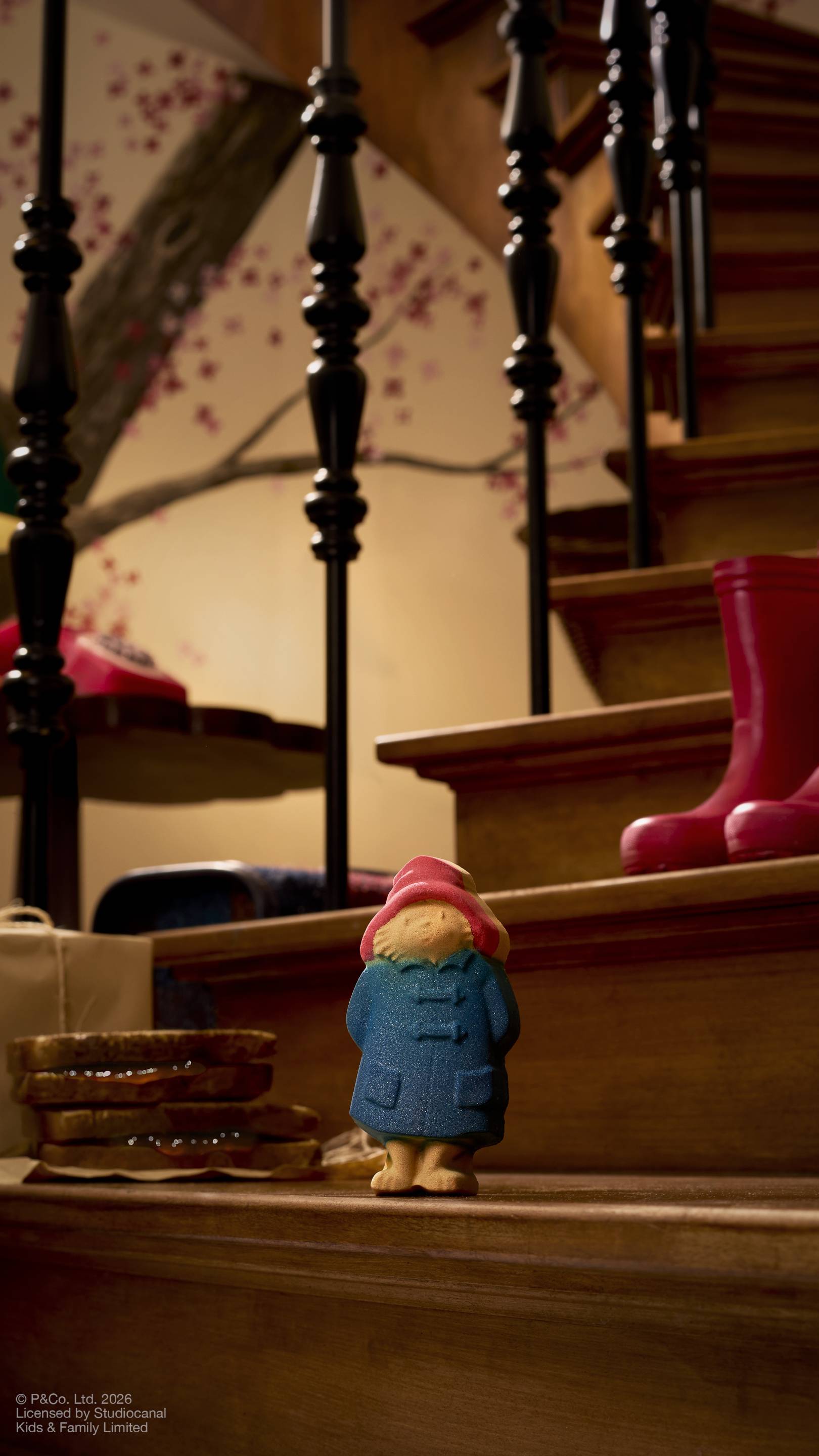 A "Paddington Bear" bath bomb sits on a wooden staircase. The bear wears his iconic red hat and blue duffle coat. Behind him, red Wellington boots rest on a step, and a stack of marmalade sandwiches sits nearby
