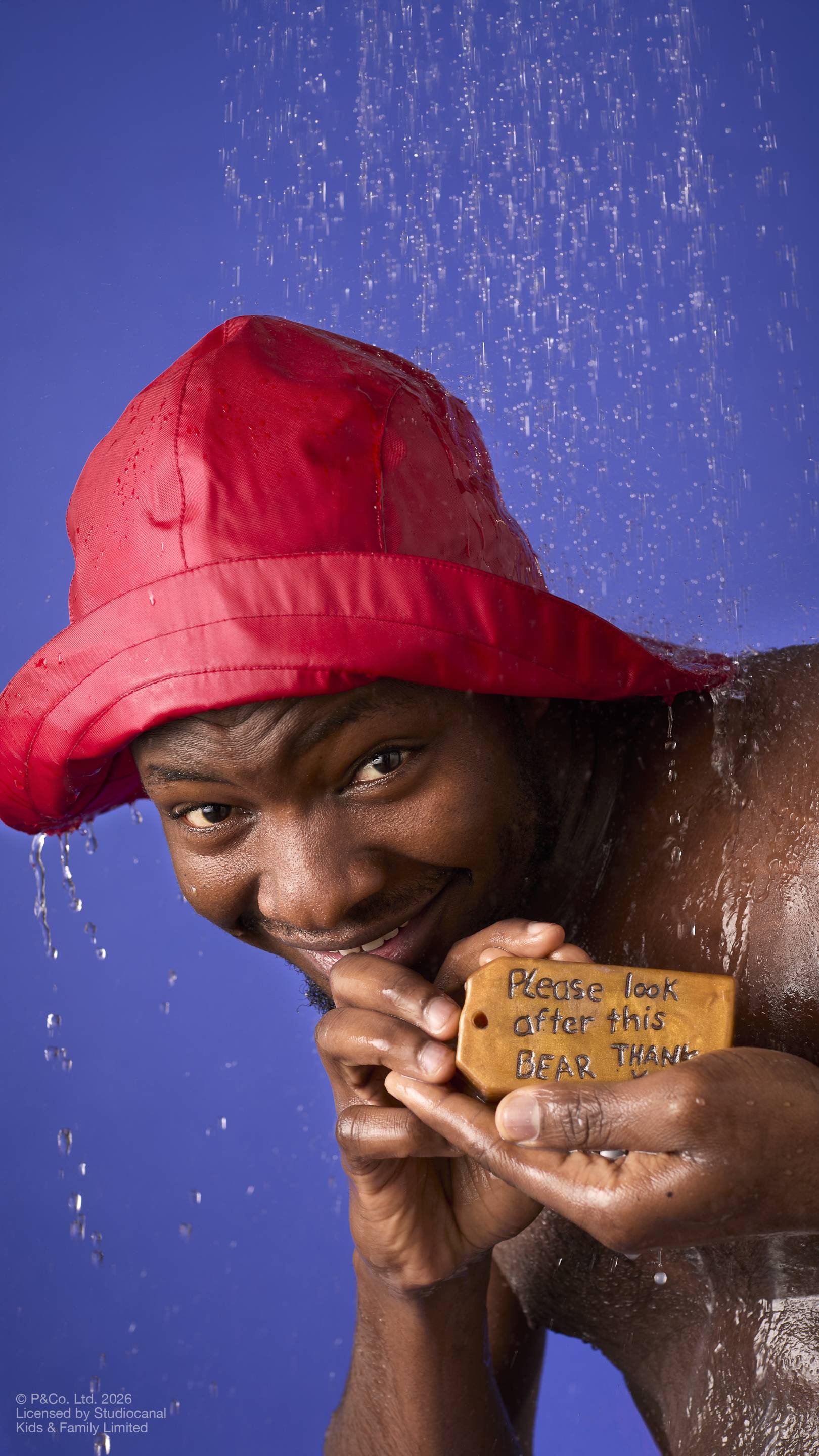 A model wearing a red rain hat stands under running shower water. They hold a brown, luggage-tag-shaped soap featuring the embossed message ‘Please look after this bear, thank you’. Droplets cascade down against a solid blue background.
