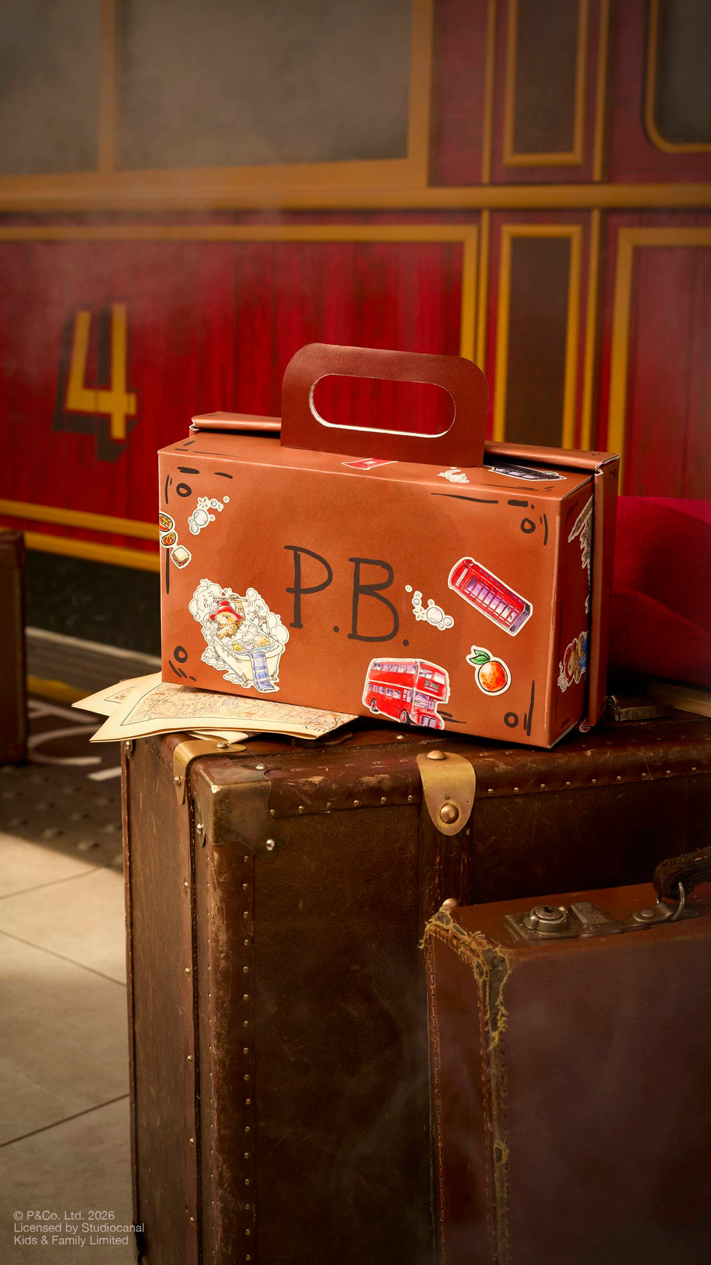 A brown, suitcase-shaped cardboard gift box with a carry handle sits on a vintage leather trunk. The box is decorated with Paddington Bear stickers, London icons like a red bus, and the initials "P.B.". A red train carriage is in the background.