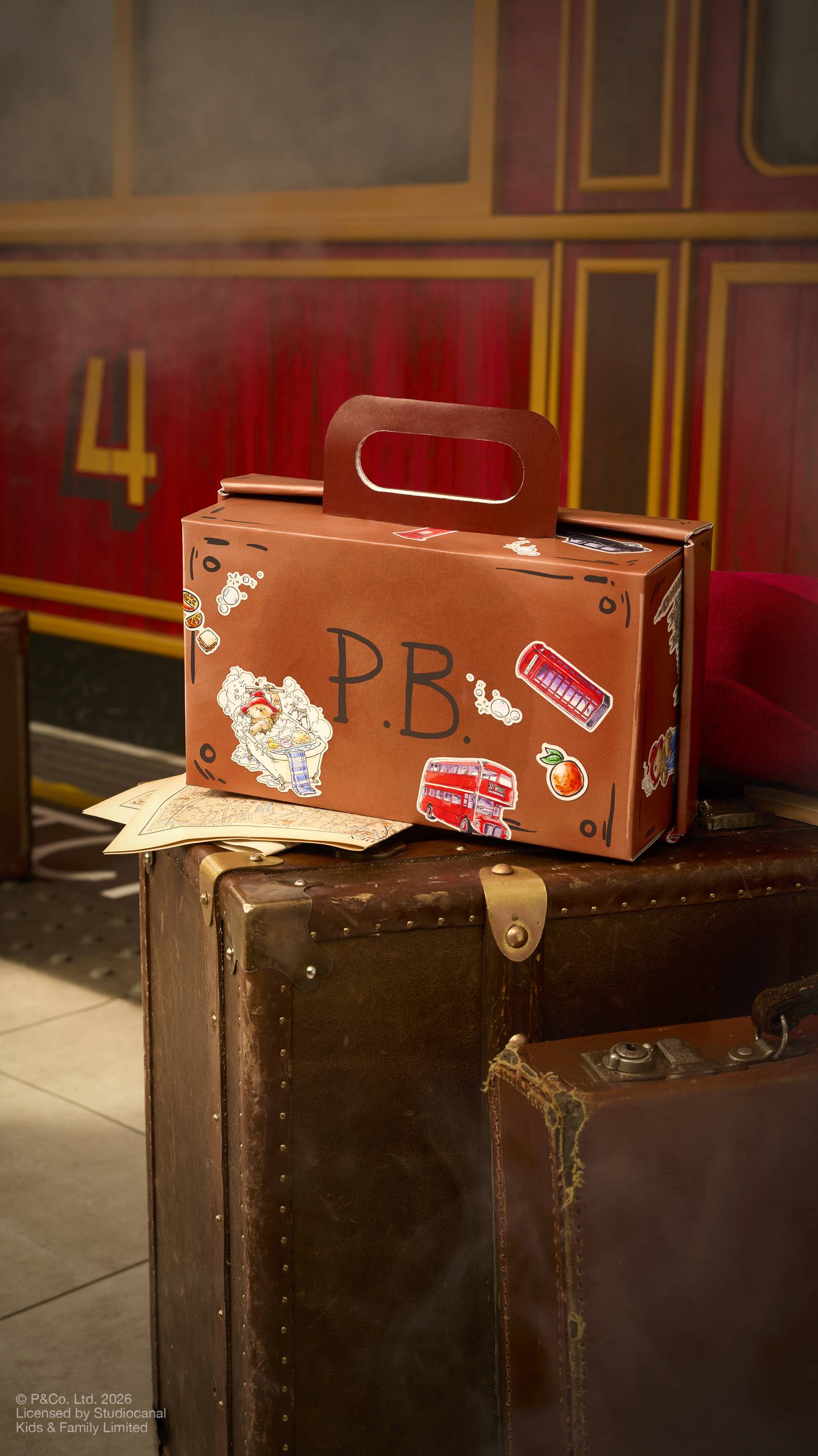 A brown, suitcase-shaped cardboard gift box with a carry handle sits on a vintage leather trunk. The box is decorated with Paddington Bear stickers, London icons like a red bus, and the initials "P.B.". A red train carriage is in the background.