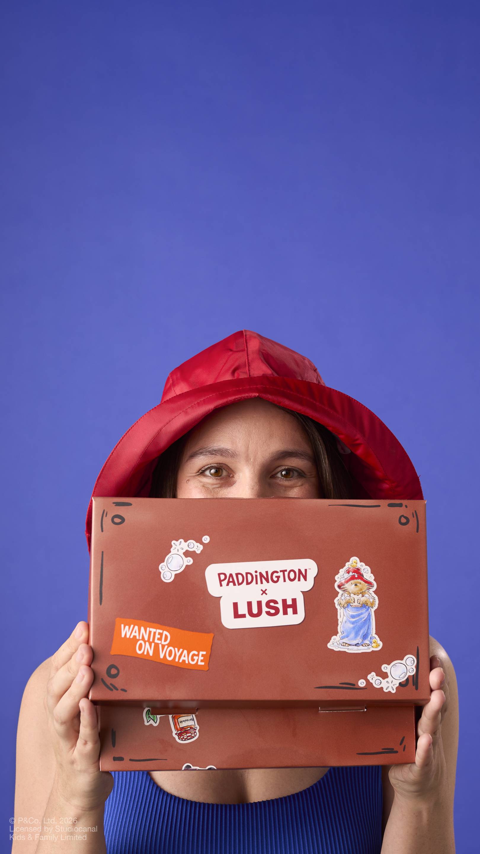 A model wearing a red rain hat peeks over a brown, suitcase-shaped gift box. The packaging features a 'PADDINGTON x LUSH' logo, a 'WANTED ON VOYAGE' sticker, and an illustration of the bear on a plain blue background.