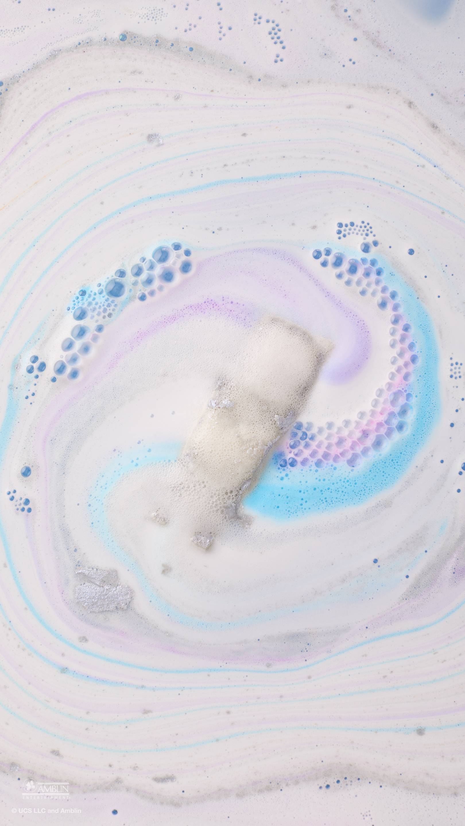 A silver, "OUTATIME" DeLorean car-shaped bath bomb completely dissolves into thick, creamy white foam. Swirls of bright blue and soft purple bubbles radiate outwards, creating a colourful, marbled pattern across the surface of the bathwater.
