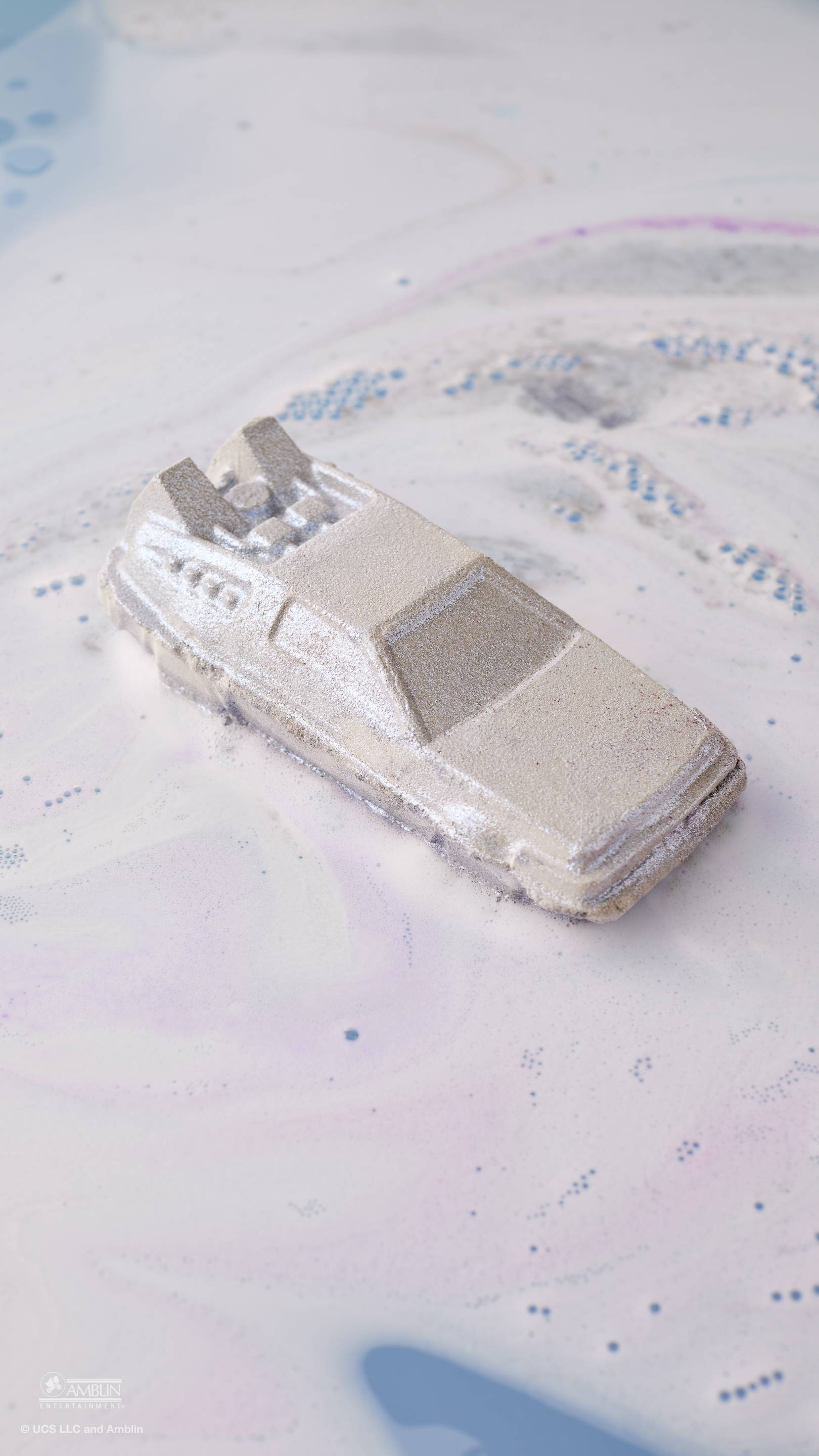 A silver, car-shaped bath bomb floats in bathwater, creating swirling clouds of white and pale purple foam. The metallic, angular car is inspired by a DeLorean and leaves a shimmering trail as it dissolves across the surface of the water.
