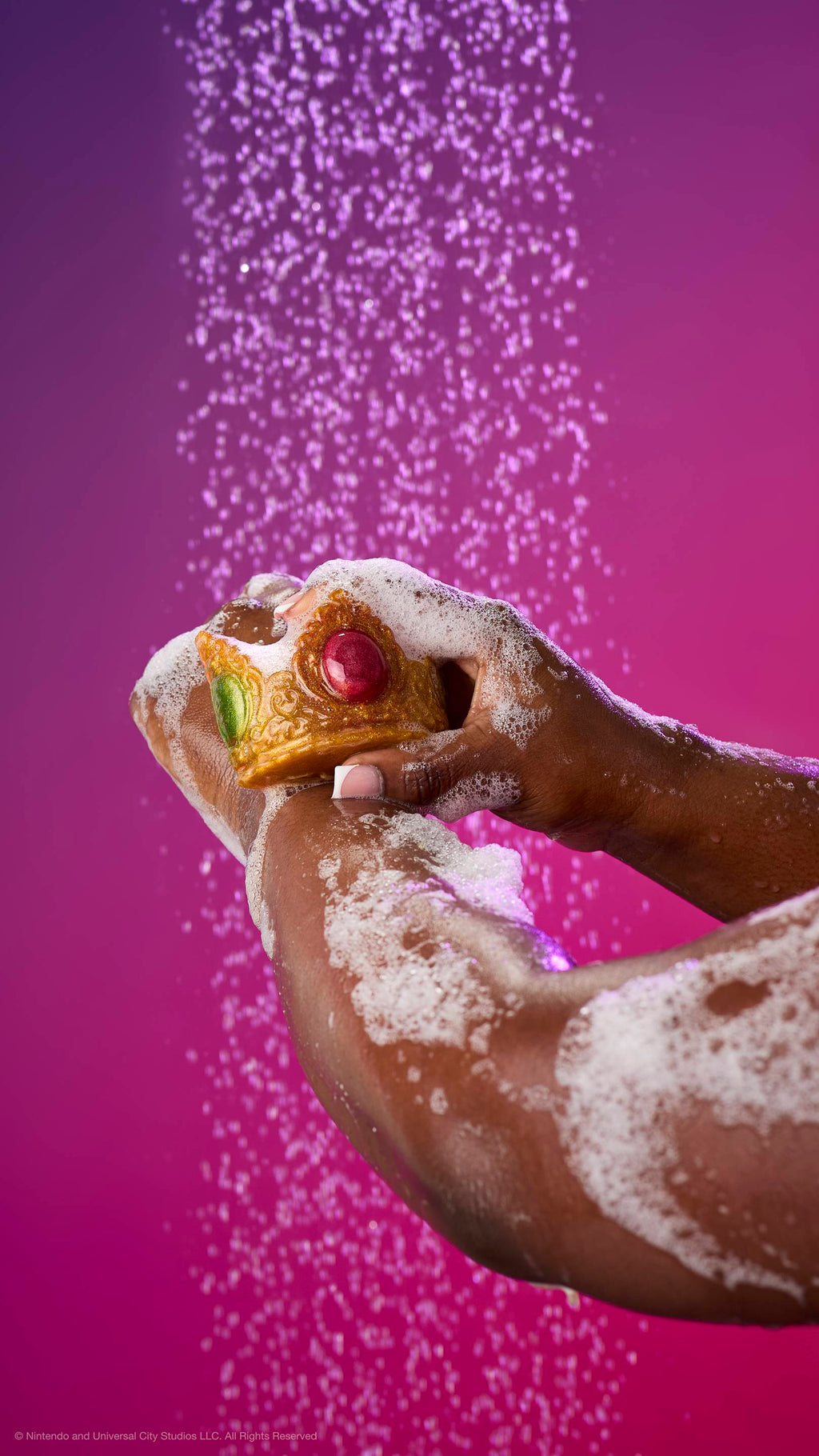 Hands lather shimmering gold Princess Peach crown soap on an arm under a shower spray. Features a large red jewel and green side jewels surrounded by white foam against a purple gradient.