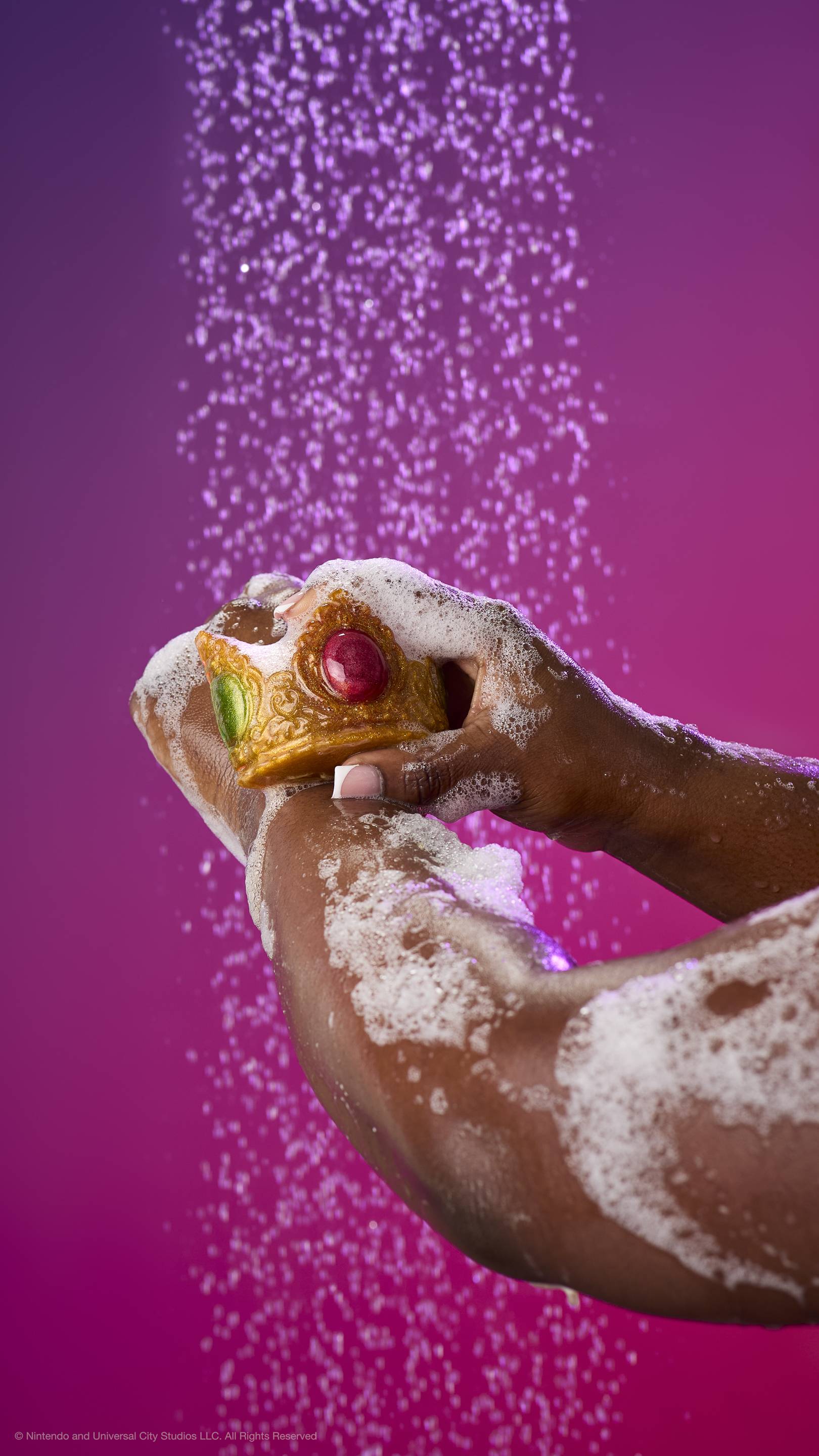 Hands lather shimmering gold Princess Peach crown soap on an arm under a shower spray. Features a large red jewel and green side jewels surrounded by white foam against a purple gradient.