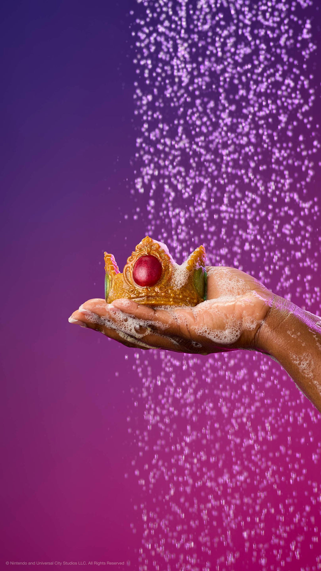 Shimmering gold Princess Peach crown soap with a large red jewel held in a soapy palm under a shower spray. Against a purple-magenta gradient with plastic-free glitter.