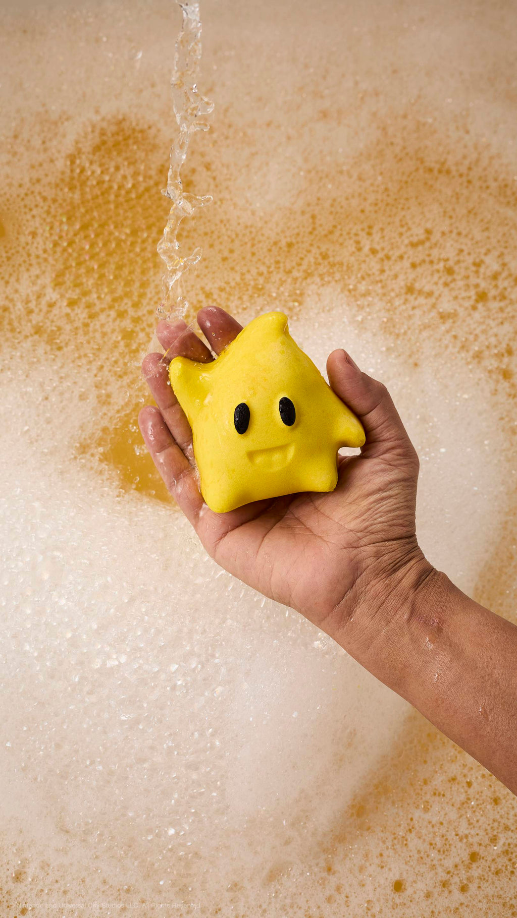 A hand holds a yellow star-shaped Luma bubble bar with black eyes and a smile under running water. The bathtub is filled with thick white foam and golden-toned bubbles.