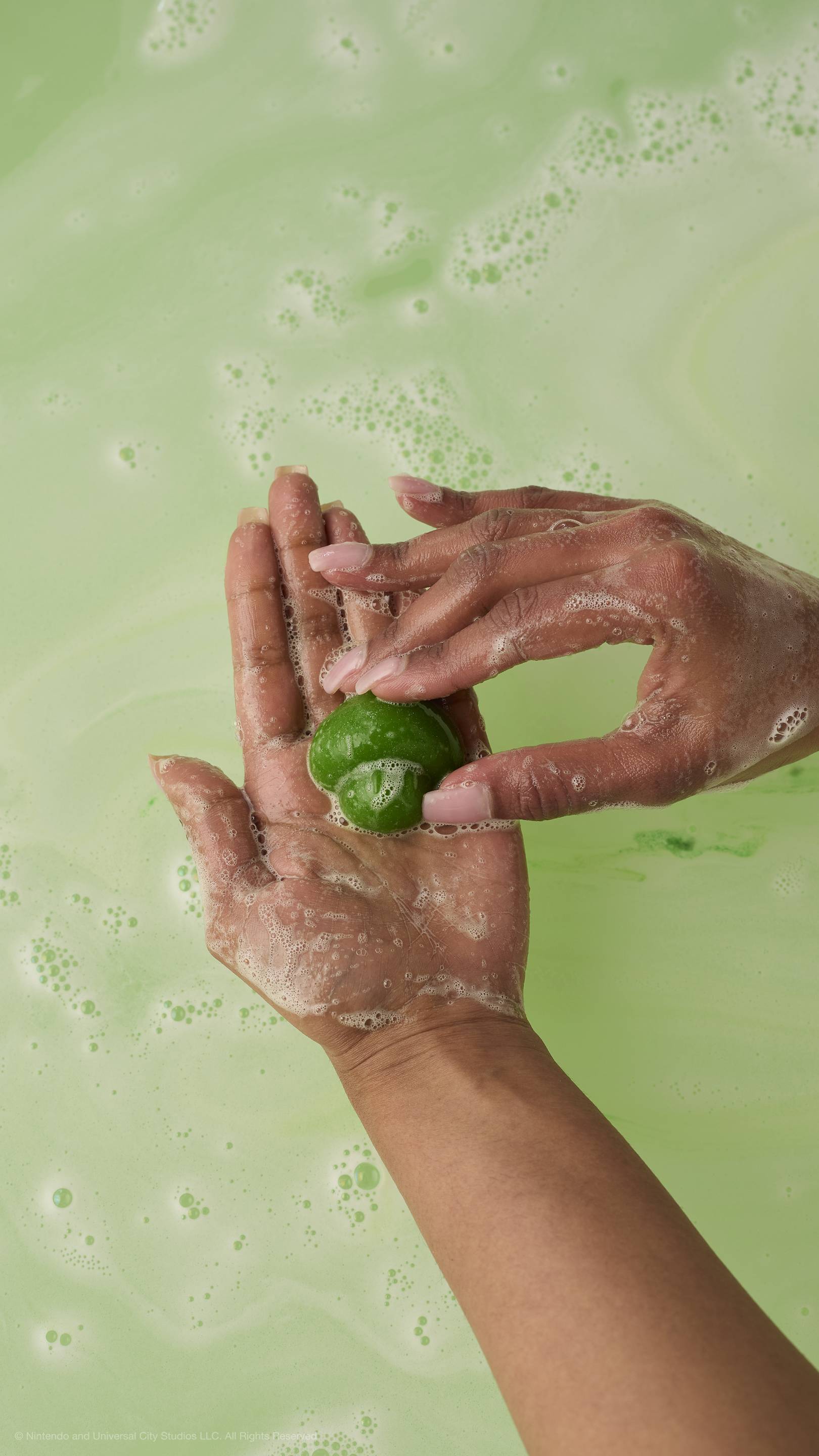 Hands hold a small, green mushroom-shaped soap over frothy, light-green bathwater.