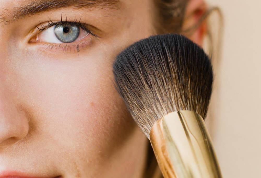A close-up of a person using People Powder, a large, dome-shaped powder brush with tapered brown bristles, on their cheekbone.