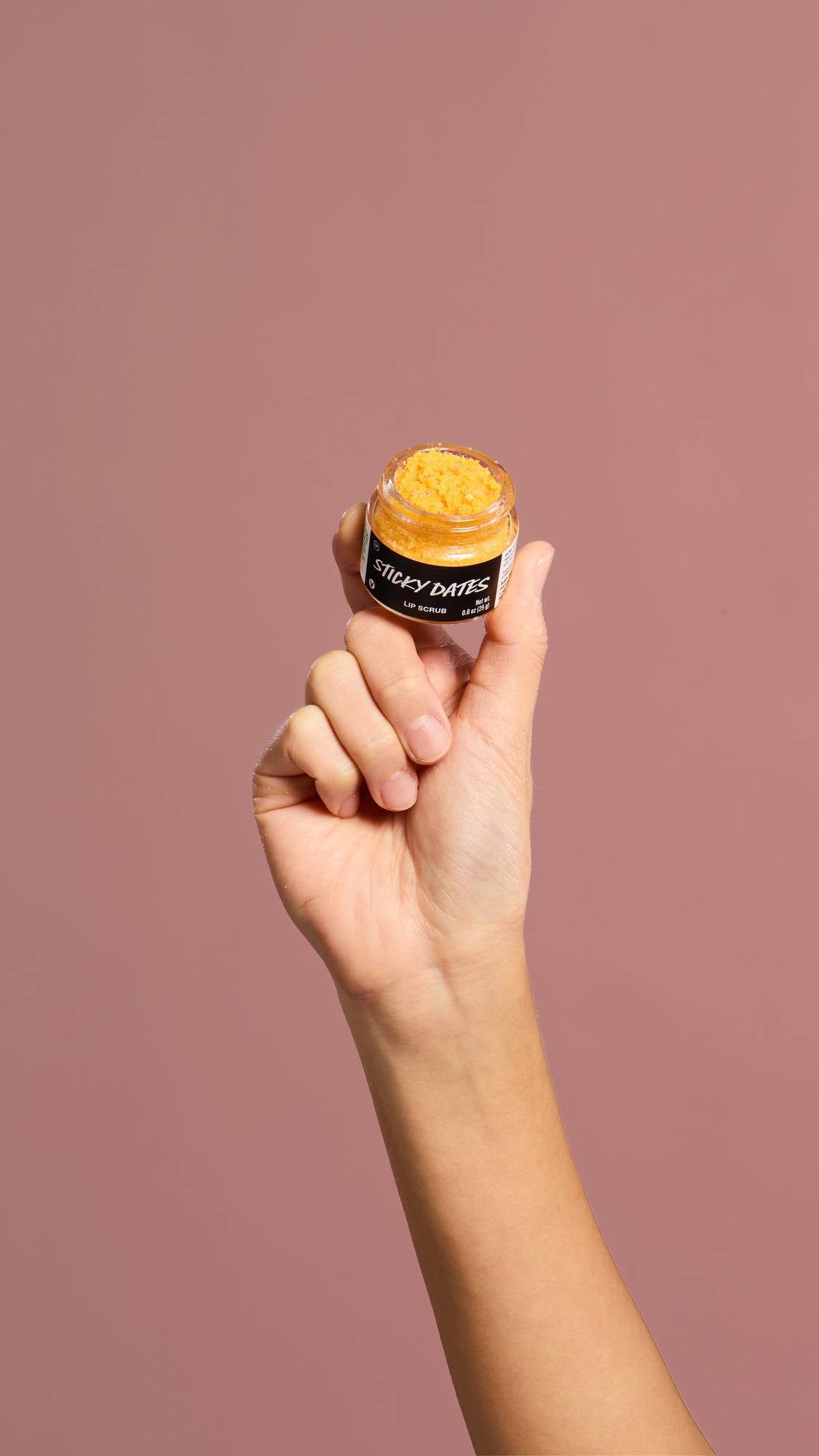 A hand holds a small open pot of "Sticky Dates" Lip Scrub against a dusty pink background. The clear glass jar contains a bright orange, granular sugar scrub.