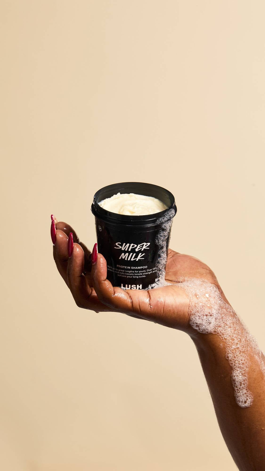 A hand with long red fingernails holds an open pot of "Super Milk" Protein Shampoo. Soapy bubbles run down the side of the black container and onto the hand, highlighting the creamy texture of the product inside against a beige background.