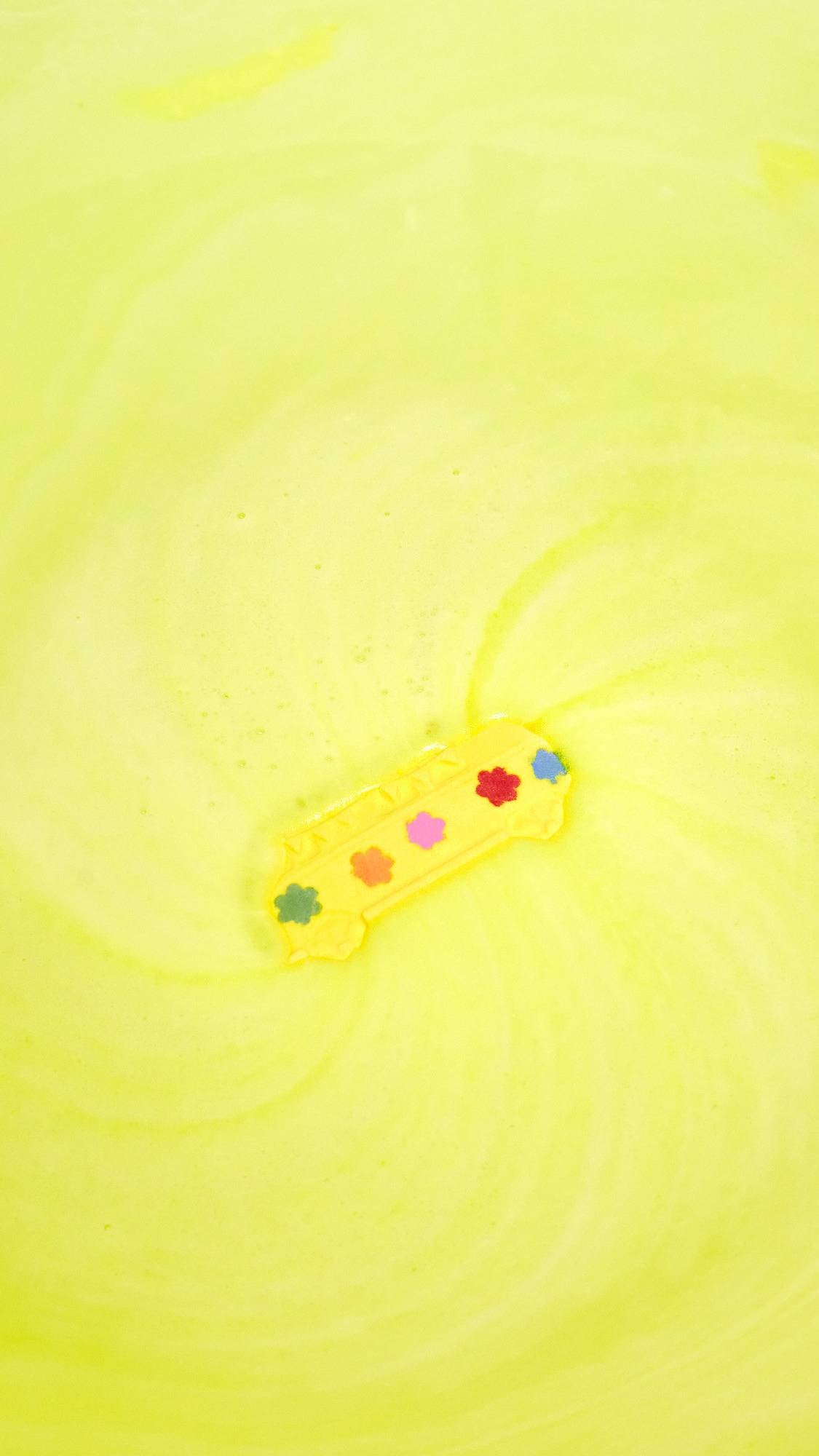 The image shows a yellow, bus-shaped product floating on the surface of a pale yellow, foamy bath. Swirling patterns radiate outwards from the bus, which has colourful embossed spots on its side.