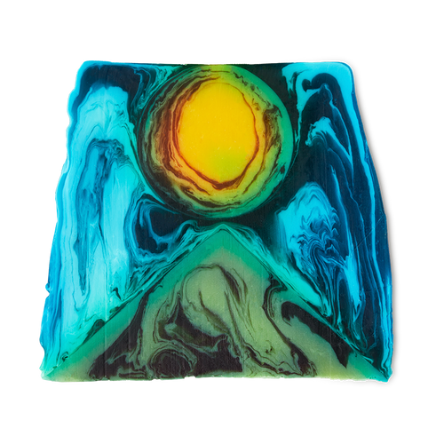 Alban Hefin is a thick, off-square slice of soap. An abstract design shows a green, pyramid-like shape at the bottom with a bright yellow, sun-like circle, surrounded by blue swirls.