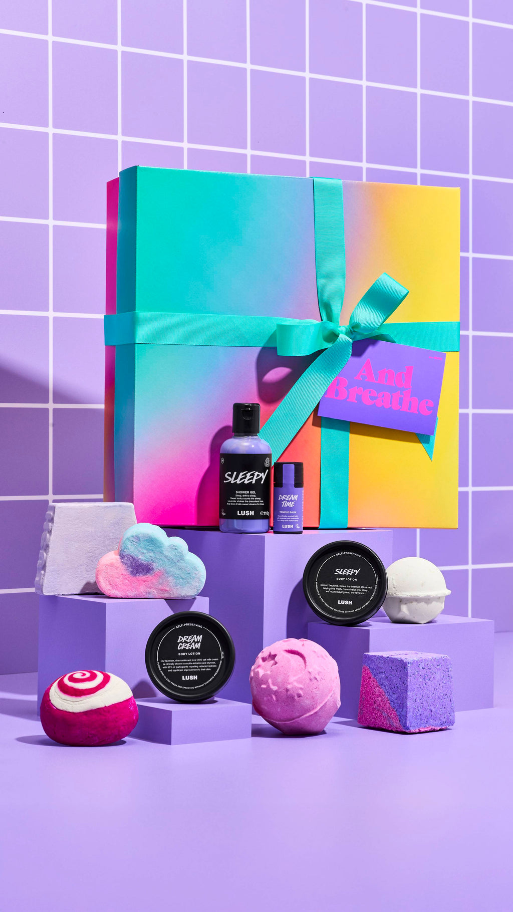 The image shows the colourful And Breathe Lush gift box sitting on a pastel purple platform on a purple-tiled background. The ten included Lush products surround the box on separate smaller platforms.