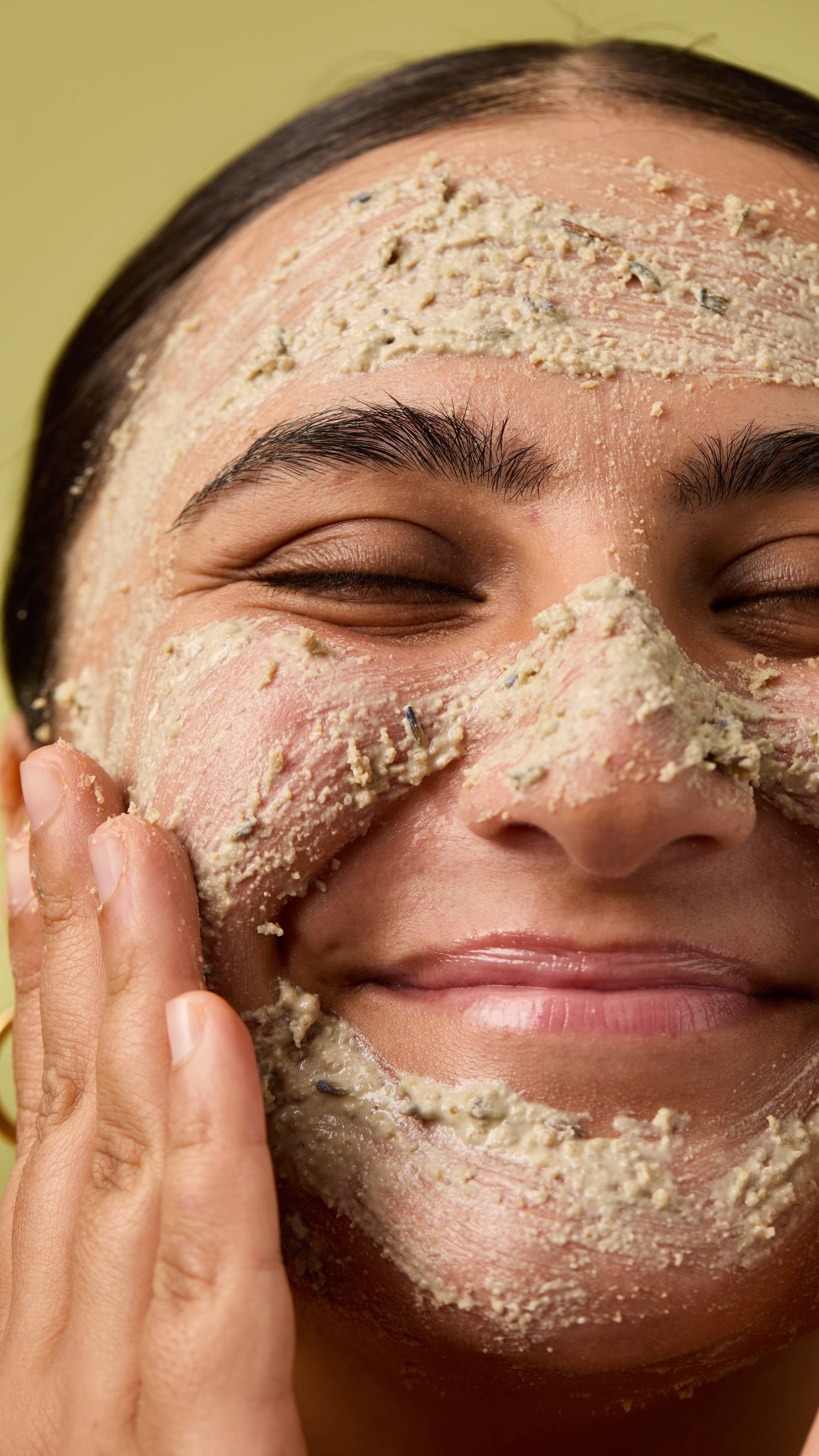 A close-up of a smiling person with eyes closed, massaging a textured, pale "Angels On Bare Skin" Lush face mask containing bits of botanical ingredients all over their face, against a muted green background.