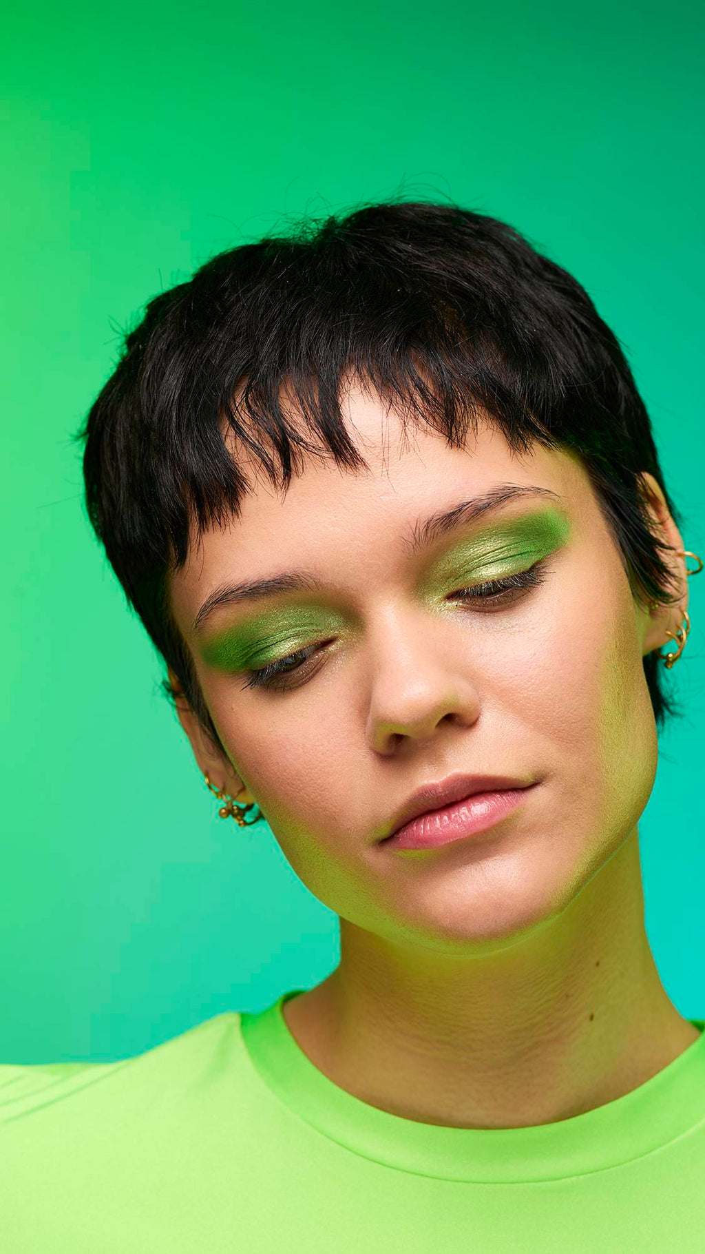 The image shows a close-up of the model's face on a pale blue-to-green gradient background. The model's make-up is done with the focal point being a subtle, jade-green, shimmering eyeshadow made by the Aurora Glitter Crumble.