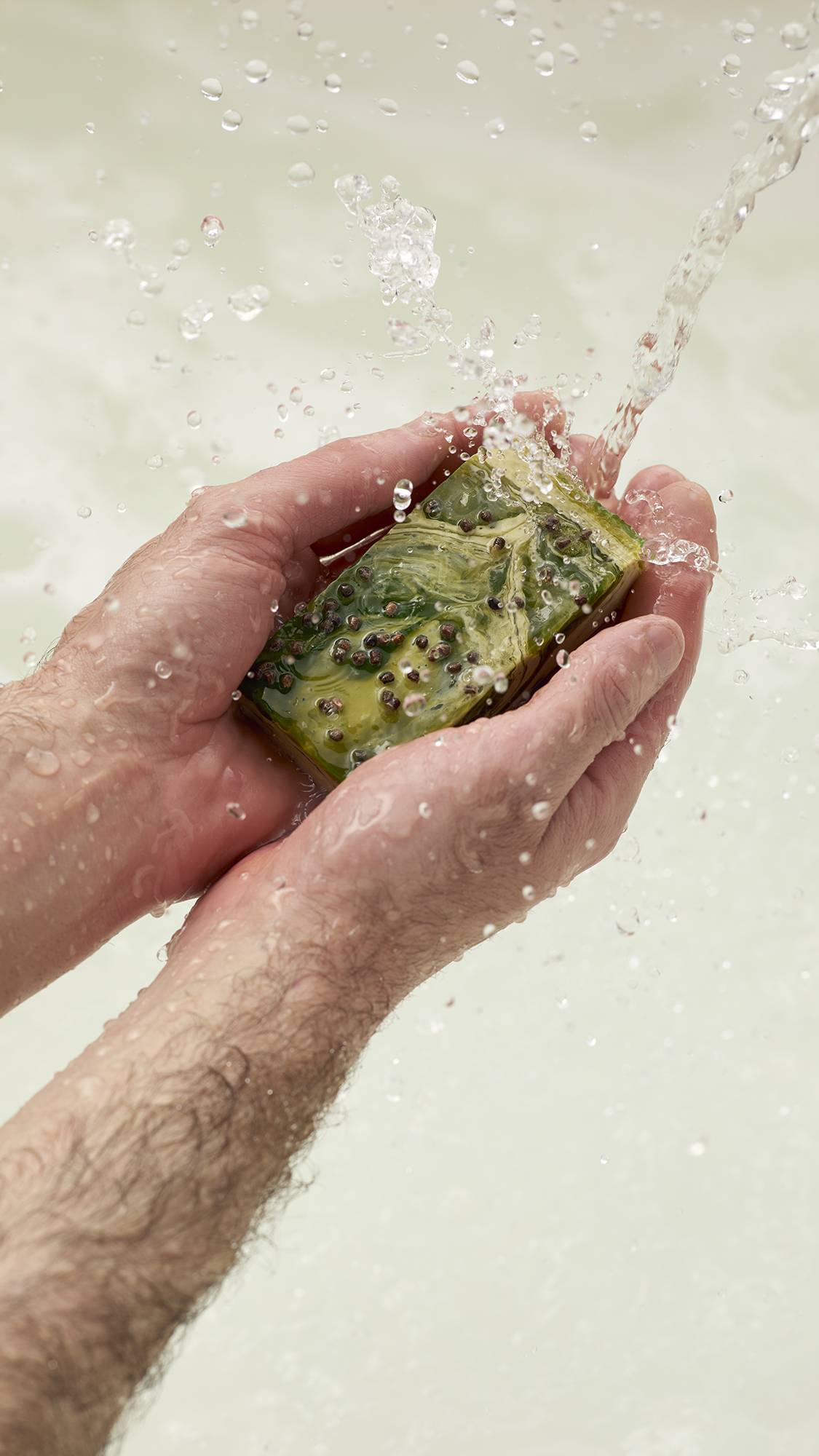 Hairy hands hold the marbled green and gold Baklava handmade soap under a splashing stream of clear water. The soap has seeds on top and is held against a soft, light neutral background.