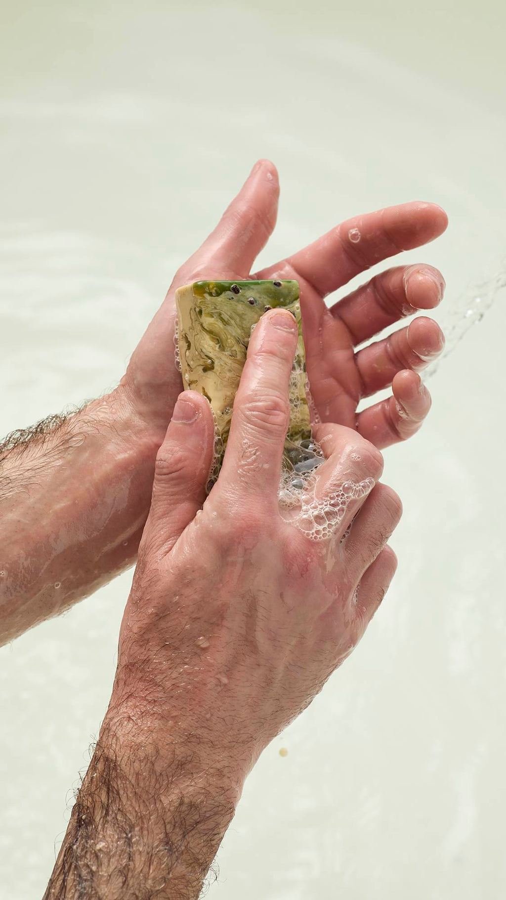 Hands lather the green and cream marbled Baklava soap, creating white bubbles. The soap features dark seeds on its surface. Set against a soft white background.