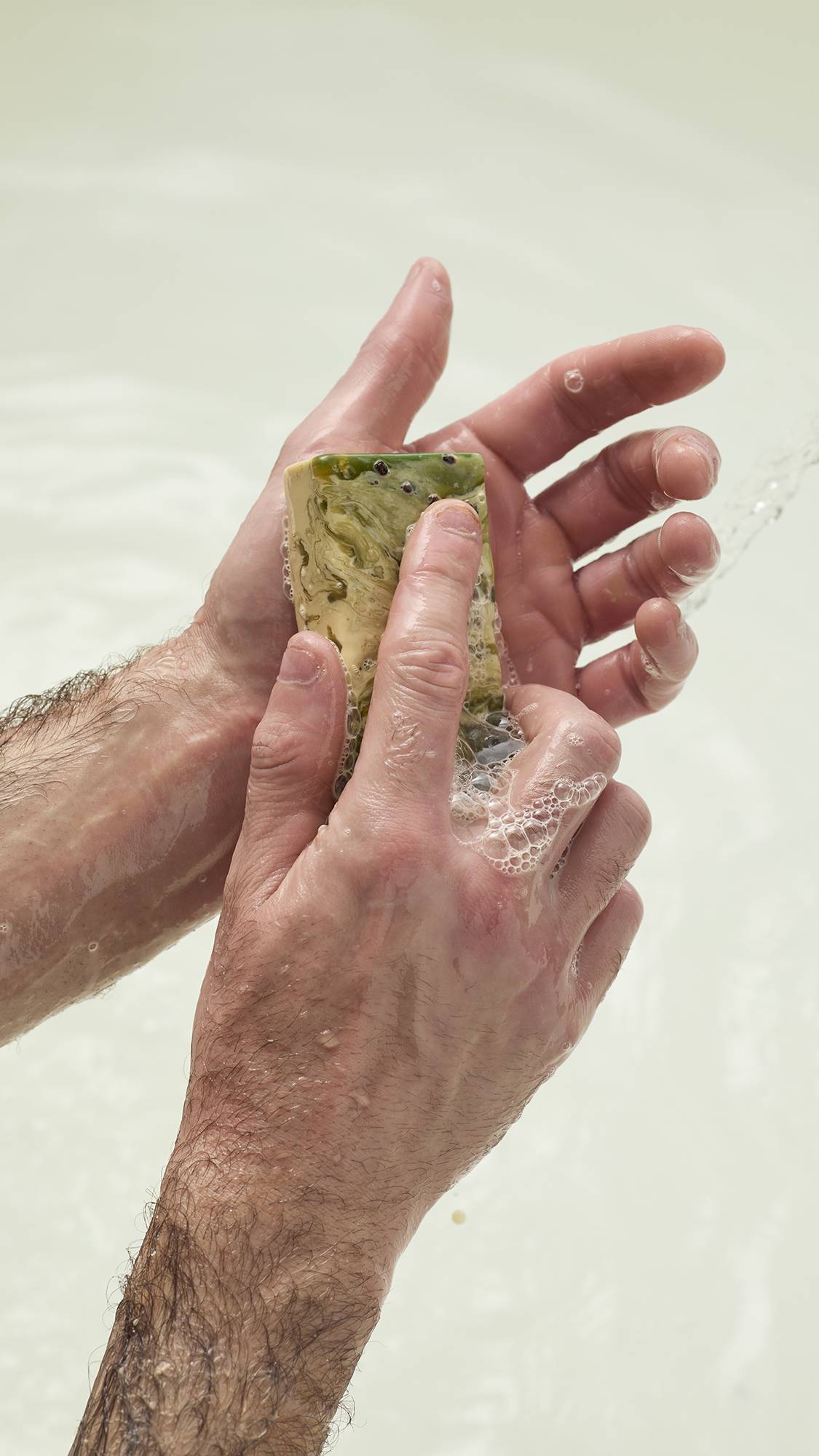 Hands lather the green and cream marbled Baklava soap, creating white bubbles. The soap features dark seeds on its surface. Set against a soft white background.