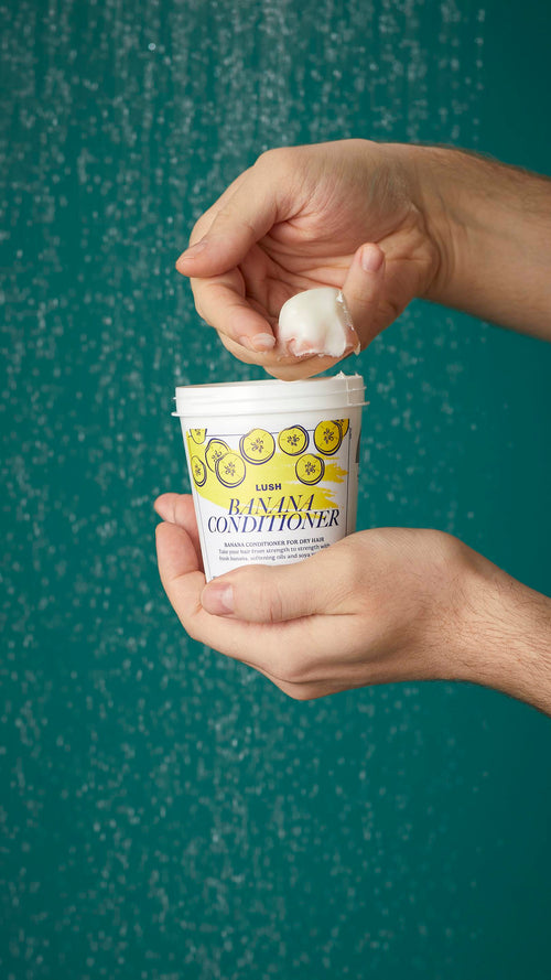 The image shows the model's arms stretched into the centre holding the white, LUSH Version-themed packed pot of the Banana conditioner. One of their hands is scooping the product out. There is a teal background.