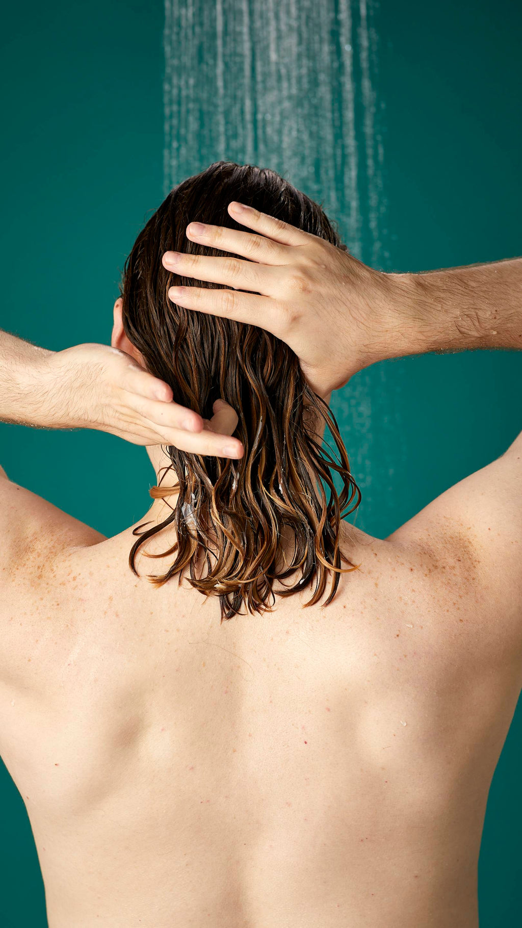The image shows the model facing away from the camera while standing under running shower water. Their hair is wet and has been shampooed already. They are now running the Banana conditioner through the lengths and ends of their hair.