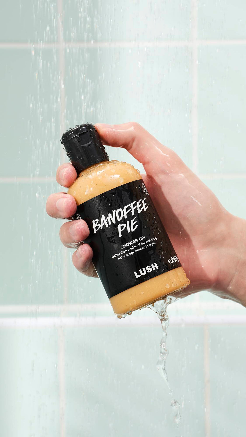 A hand holds a bottle of Lush Banoffee Pie shower gel under a stream of water in a shower. The clear bottle reveals the creamy yellow gel inside, and the black label features white text. Mint green tiles are in the background.