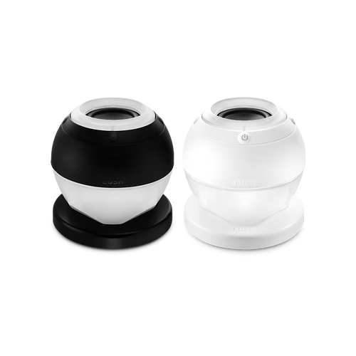 Bath Bot. Two slick, bath bomb-shaped speaker gadgets with a flat base and modern design. One is fully white and the other is black and white. There is an embossed power button visible and the word "LUSH".