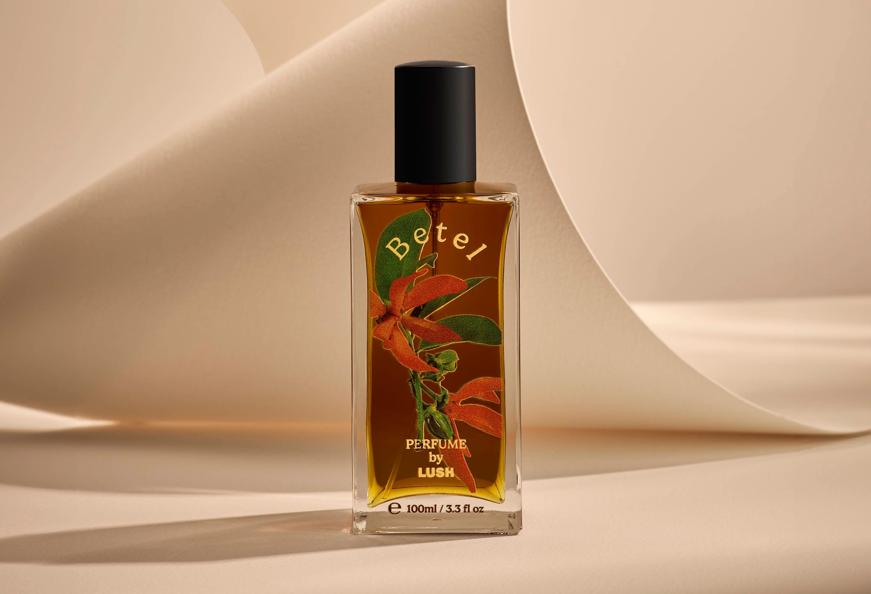 The image shows the Betel glass perfume bottle. It is sitting on a soft, beige background with curved, cream paper creating soft, gliding shapes behind the product.