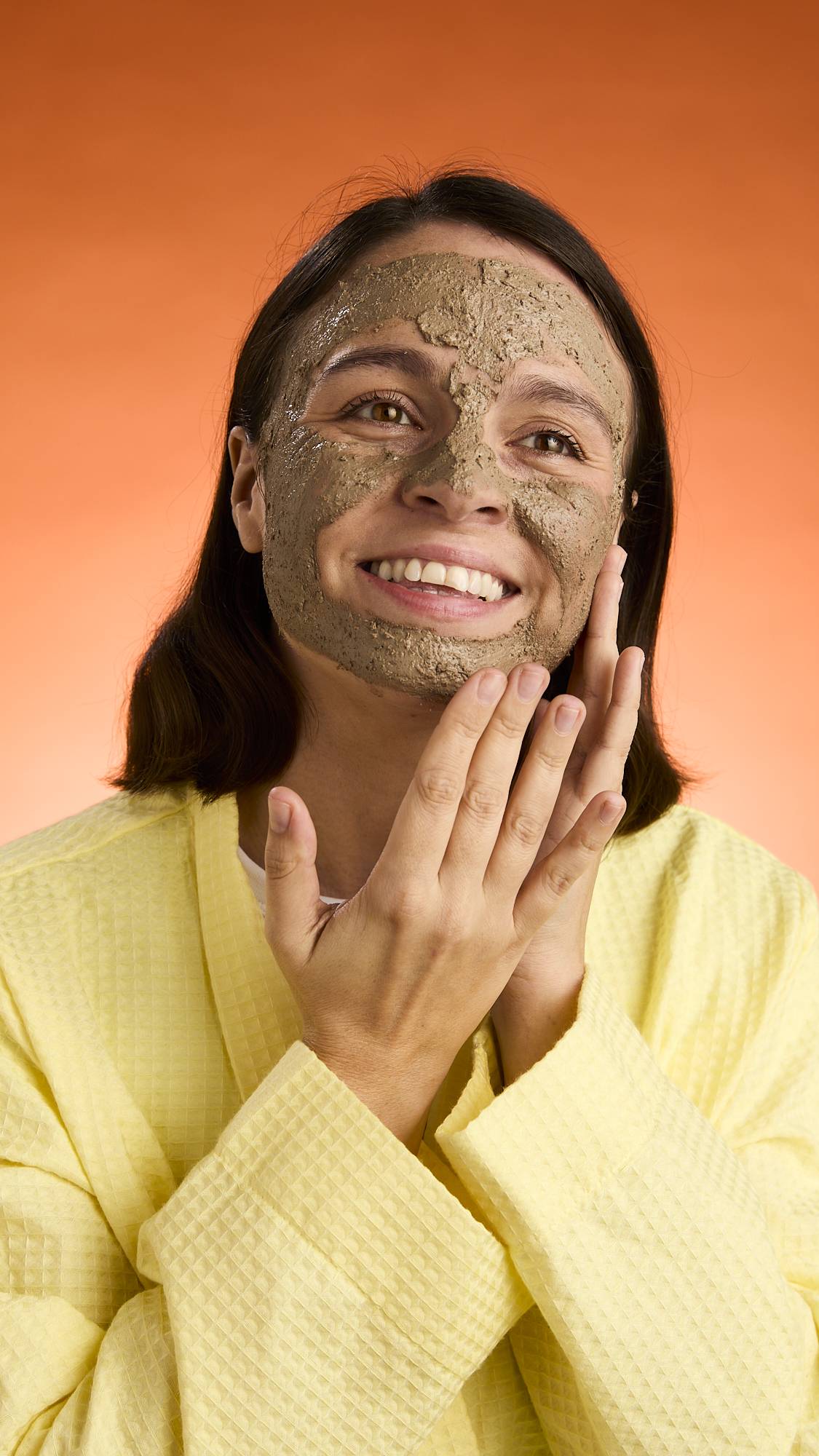 A smiling person with dark hair wears a yellow robe and touches their face, showcasing the full application and texture of the Black Tea Mochi face mask against an orange background.