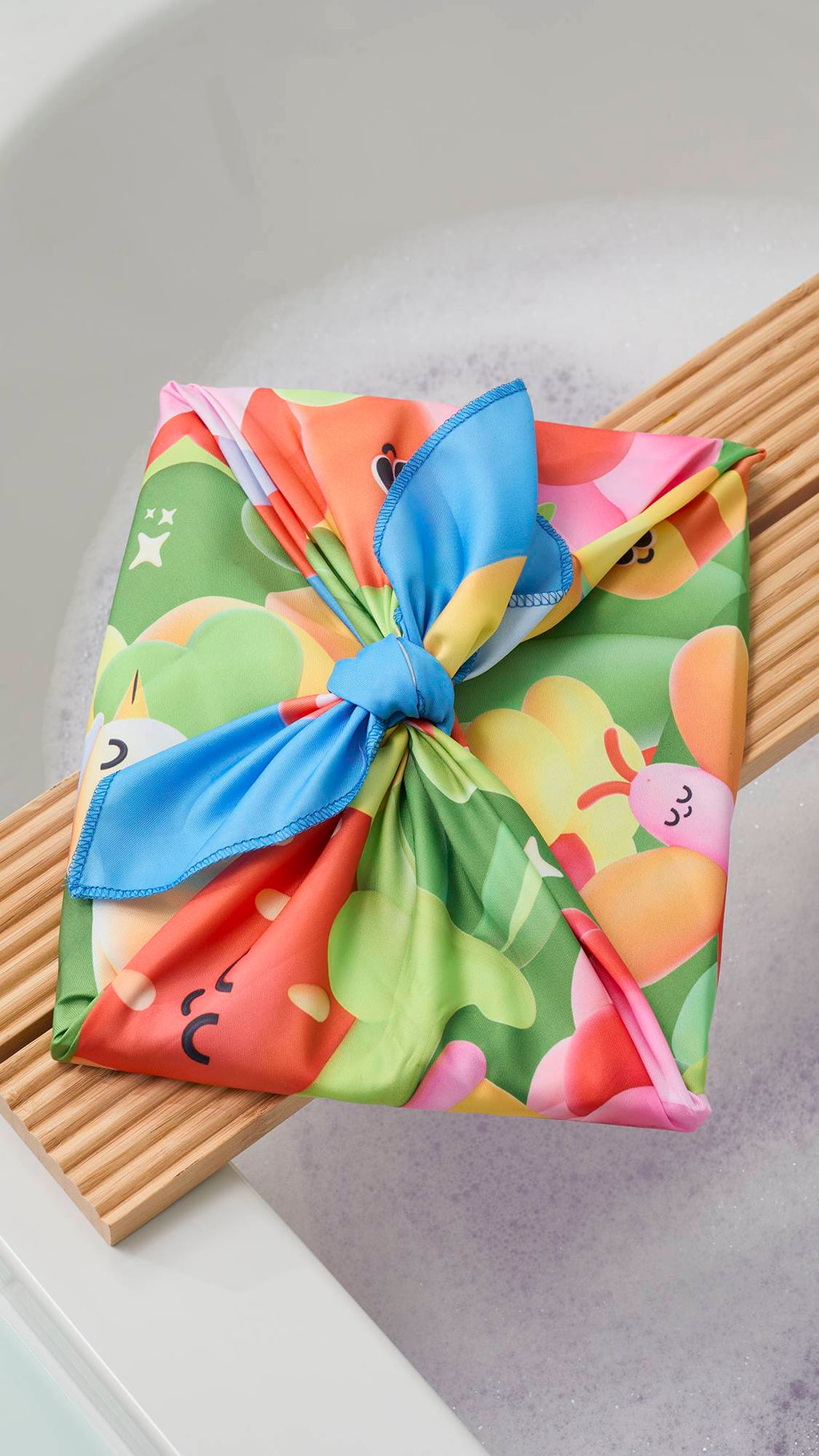 A vibrant "Blooming Lovely" fabric Knot Wrap, tied in a decorative bow, rests on a wooden bath trap over a soapy bathtub. The fabric features a colourful, 3D-style garden print with a large red strawberry and green foliage.