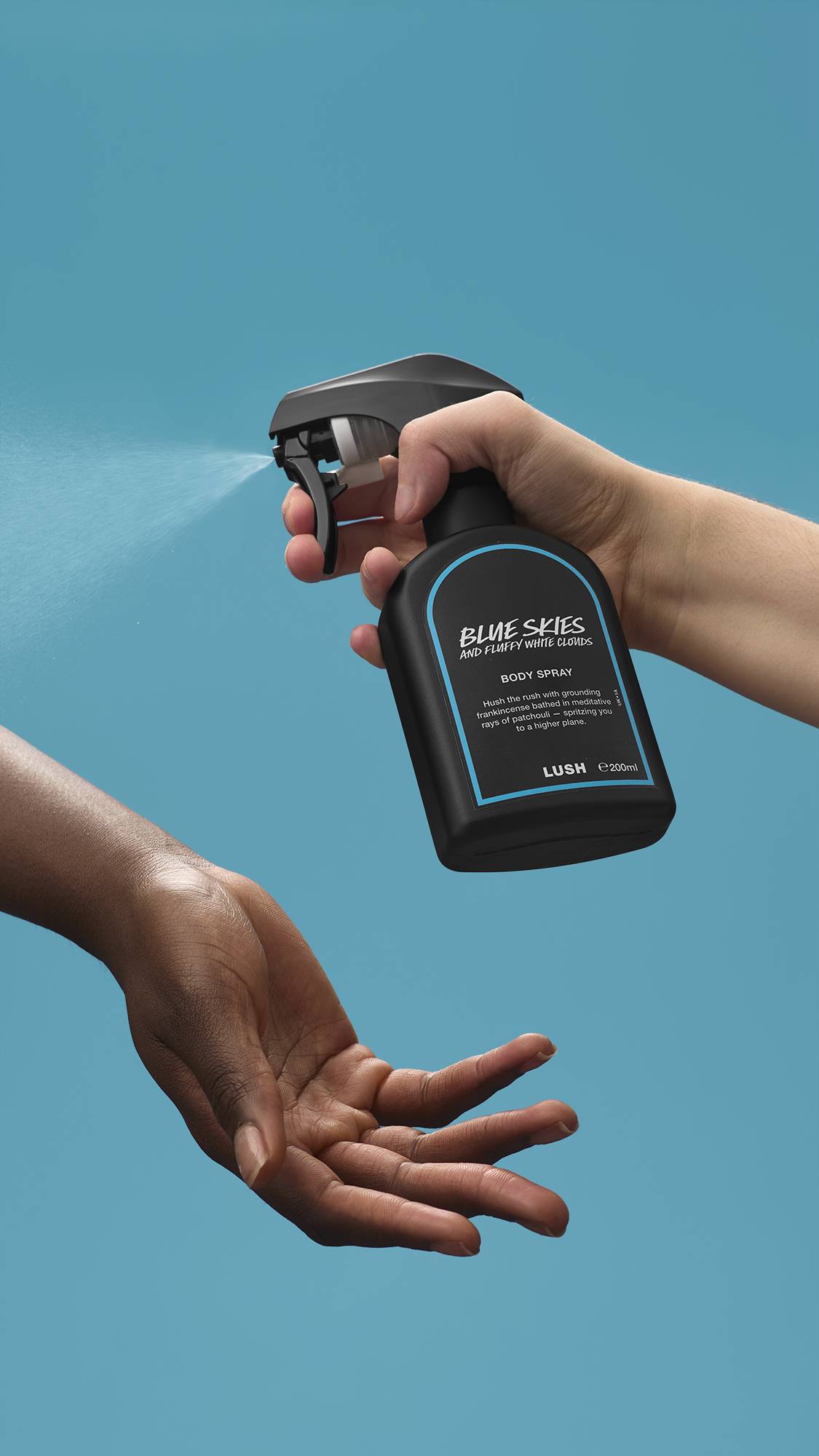 The image shows a light-toned hand spraying a bottle of LUSH 'Blue Skies and Fluffy White Clouds' body spray. A warm-toned hand is cupped below, catching the fine mist against a bright, sky-blue background.
