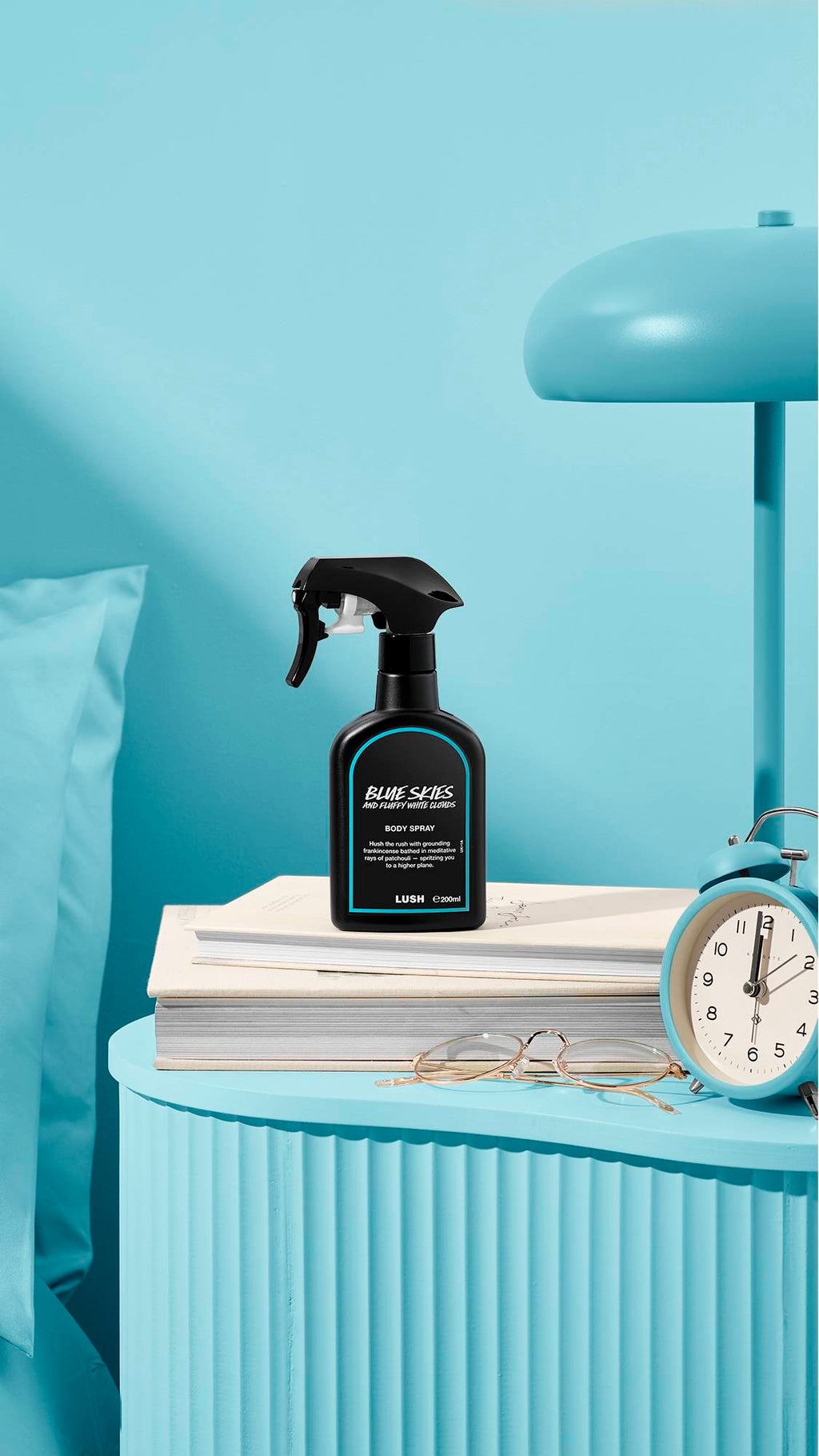 The image shows a recycled black plastic LUSH spray bottle of "Blue Skies and Fluffy White Clouds" sitting on a pile of books on a light blue bedside table. A matching lamp, alarm clock, and glasses sit nearby, next to a blue bed and wall.