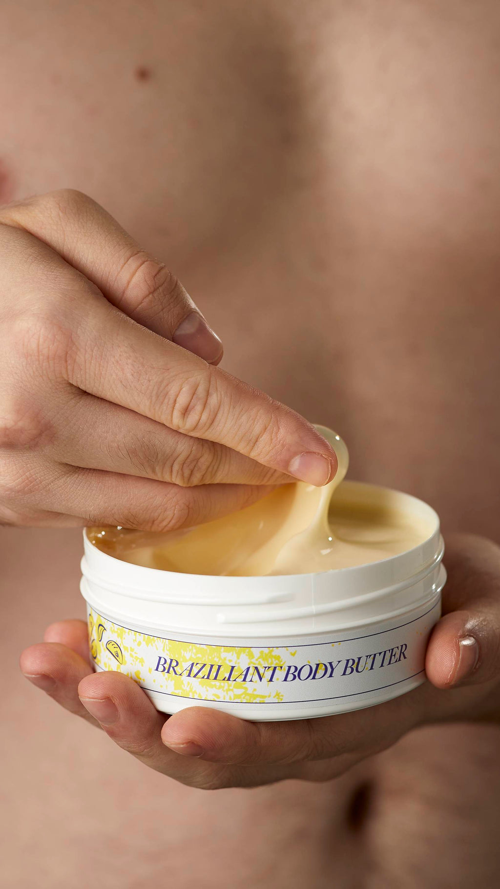 A close-up shot of the model's hands. One hand is holding the flat round, white, LUSH Version-themed packed tub of Braziliant body butter.