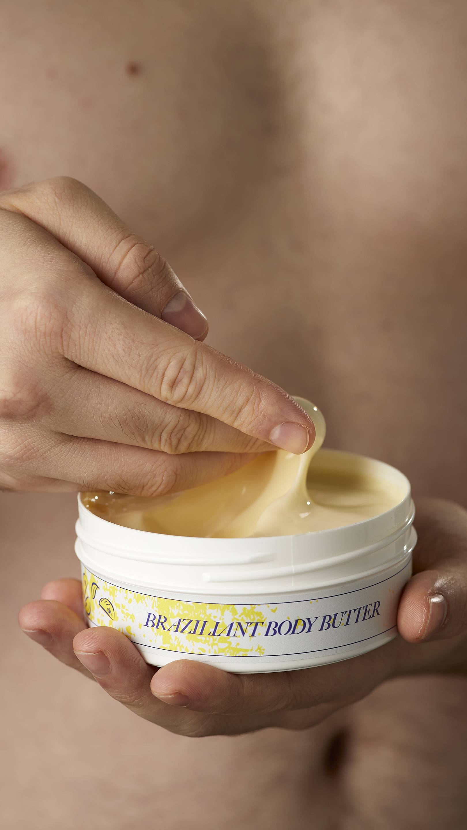 A close-up shot of the model's hands. One hand is holding the flat round, white, LUSH Version-themed packed tub of Braziliant body butter.