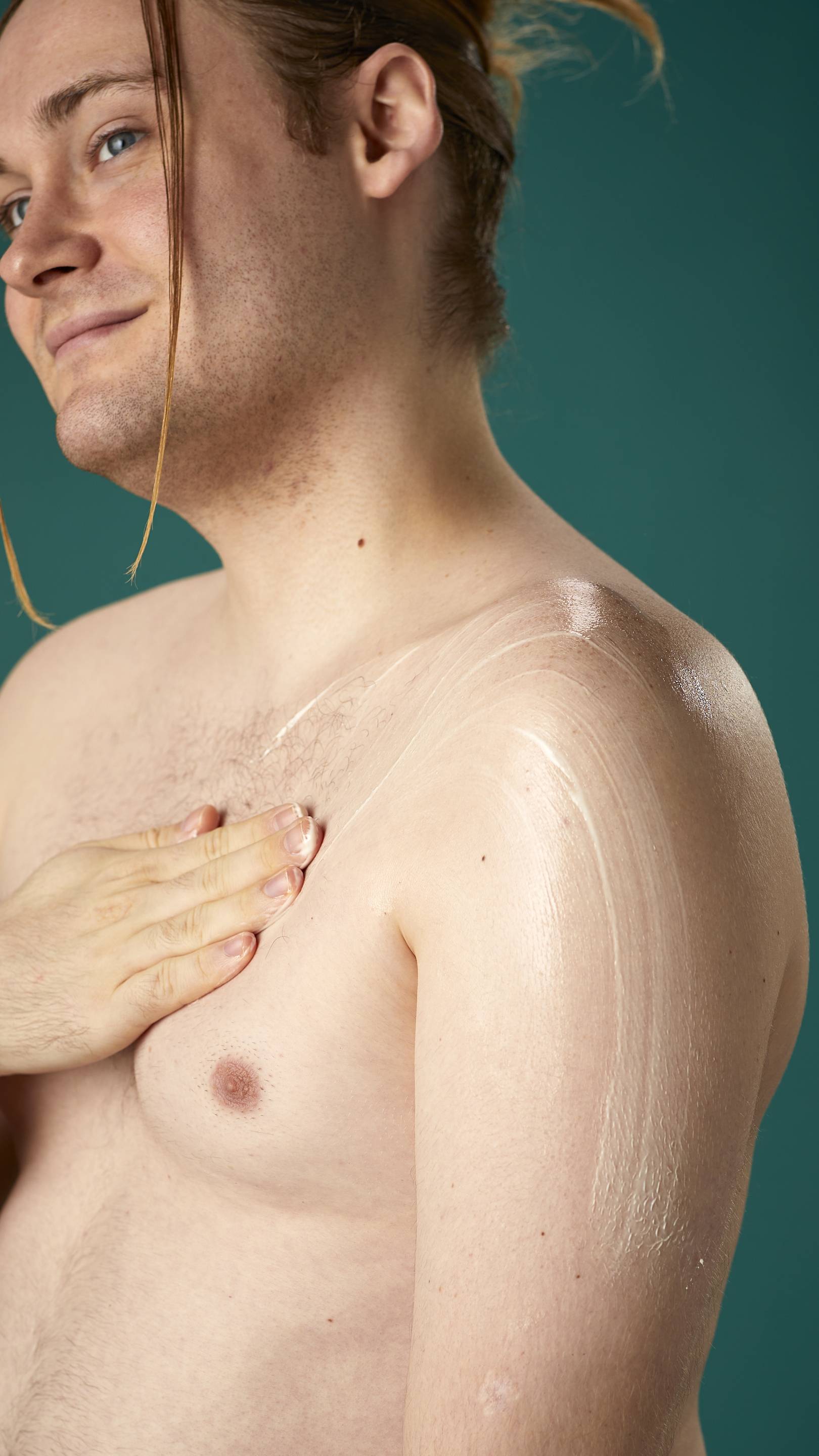 The image shows a topless model on a teal background. They are gently soothing the Braziliant body butter in circular motions over their chest and upper arm.
