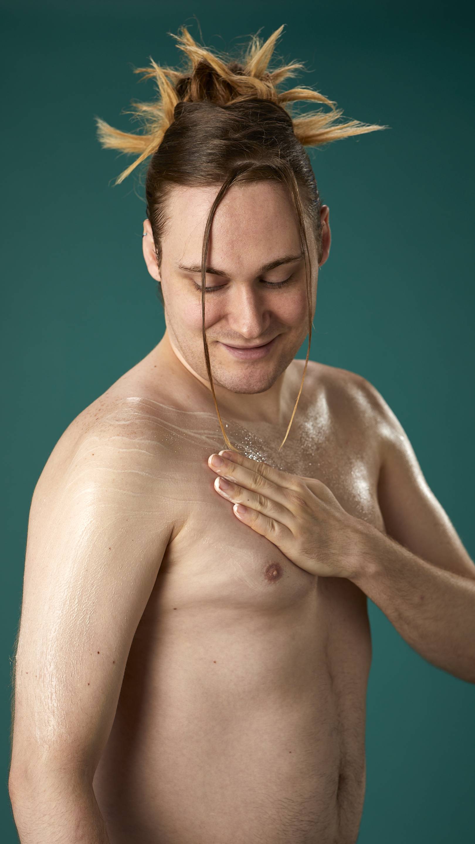 The image shows a topless model on a teal background. They are gently soothing the Braziliant body butter in circular motions over their chest and upper arm.
