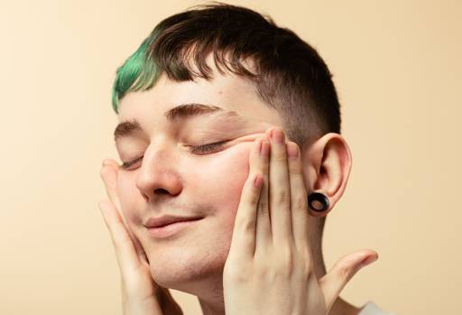 A person with short hair, partly coloured green, smiles blissfully, as they enjoy pushing their cheeks upwards with their hands.