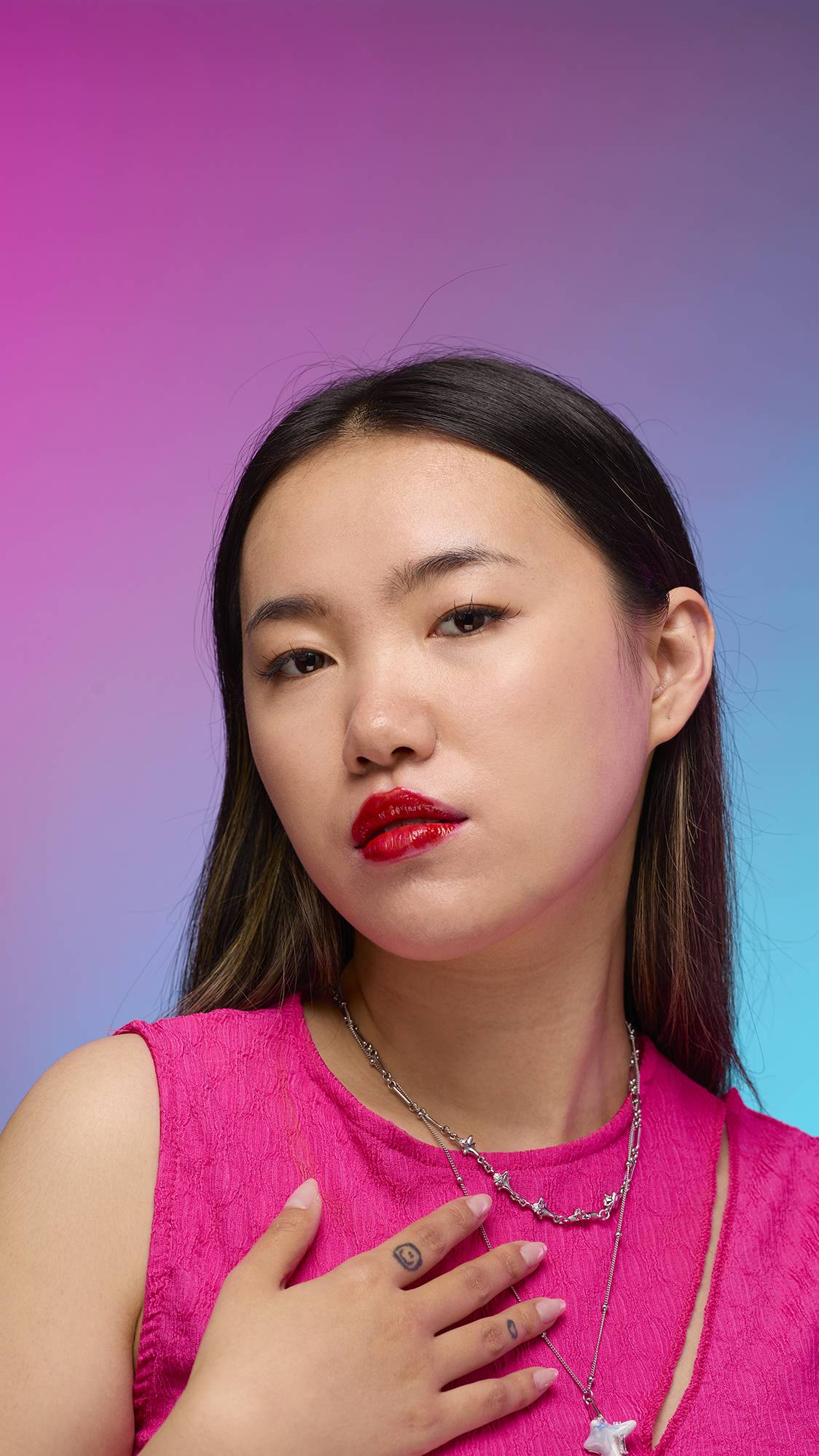 The model has long, straight dark hair and is on a pastel pink, blue and purple hue background. Their make-up has been done with the focal point being the juicy coating of cherry-red tint on their lips created by the Cherry lip jelly.